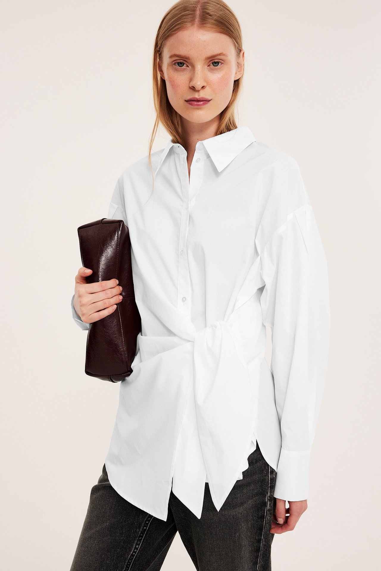 Framifrån: kvinna i vit bomullsskjorta med knytning framtill, mörka jeans, håller mörkbrun clutch.