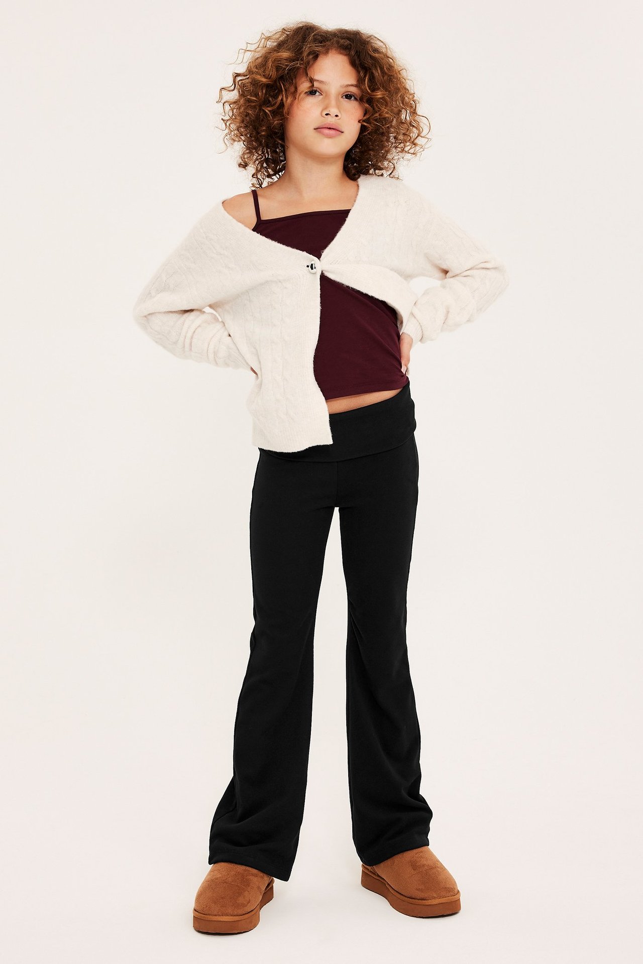 Edestä: Lapsi mustissa levenevissä leggingseissä, viininpunaisessa topissa, kermanvärinen neuletakki, ruskeat tohvelit.