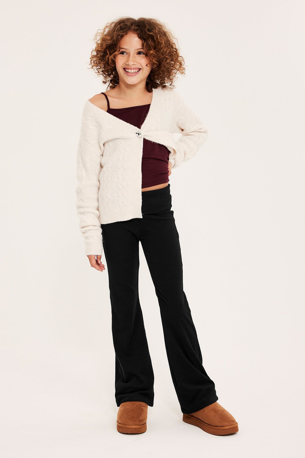 Widok z przodu: dziecko w czarnych legginsach flare, bordowym topie, kremowym kardiganie i brązowych butach.