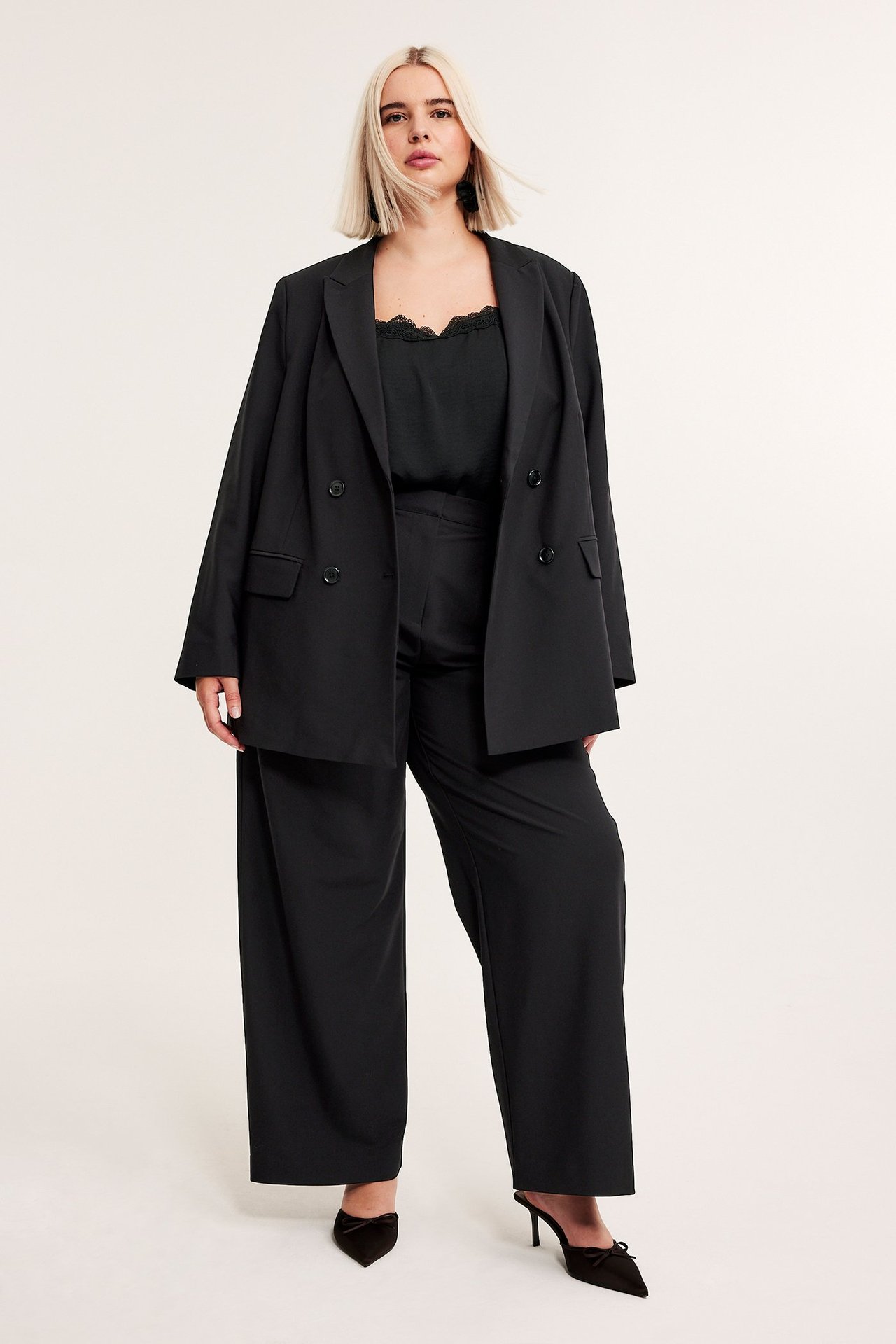 Edestäpäin, nainen mustassa kaksirivisessä bleiserissä, topissa, leveälahkeisissa housuissa ja mustissa koroissa.