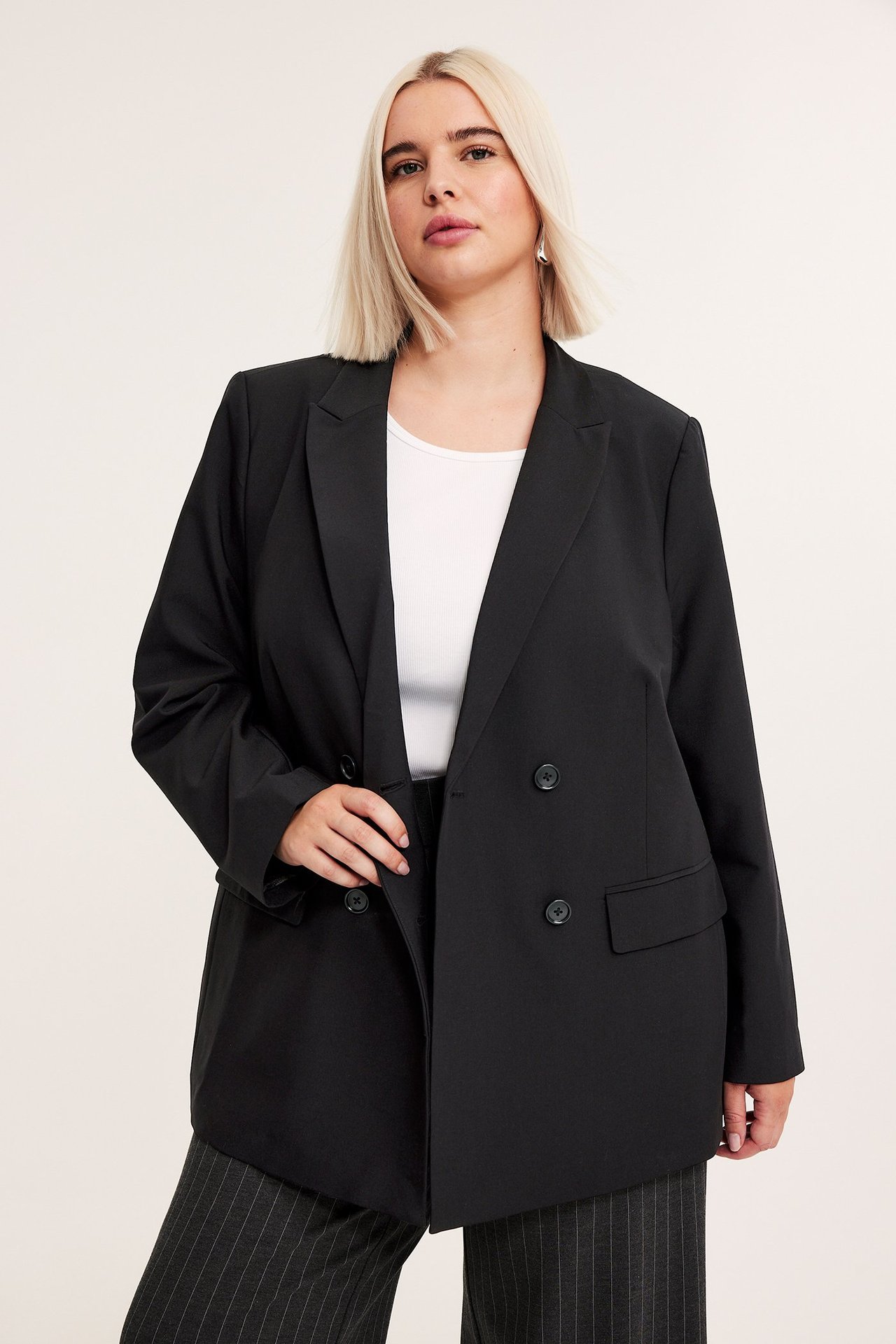 Widok z przodu: kobieta w czarnej dwurzędowej marynarce, białym topie i ciemnych spodniach w paski.