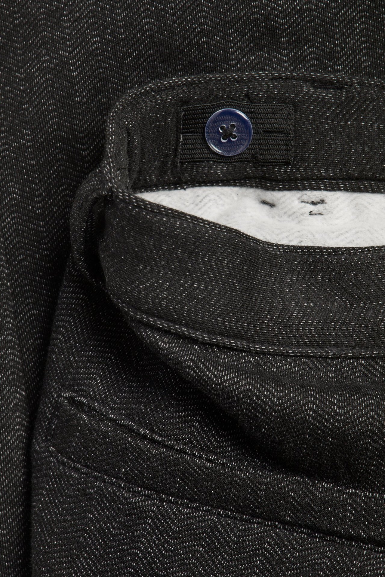 Zbliżenie ciemnoszarych spodni w jodełkę z regulowaną elastyczną talią i niebieskim guzikiem.