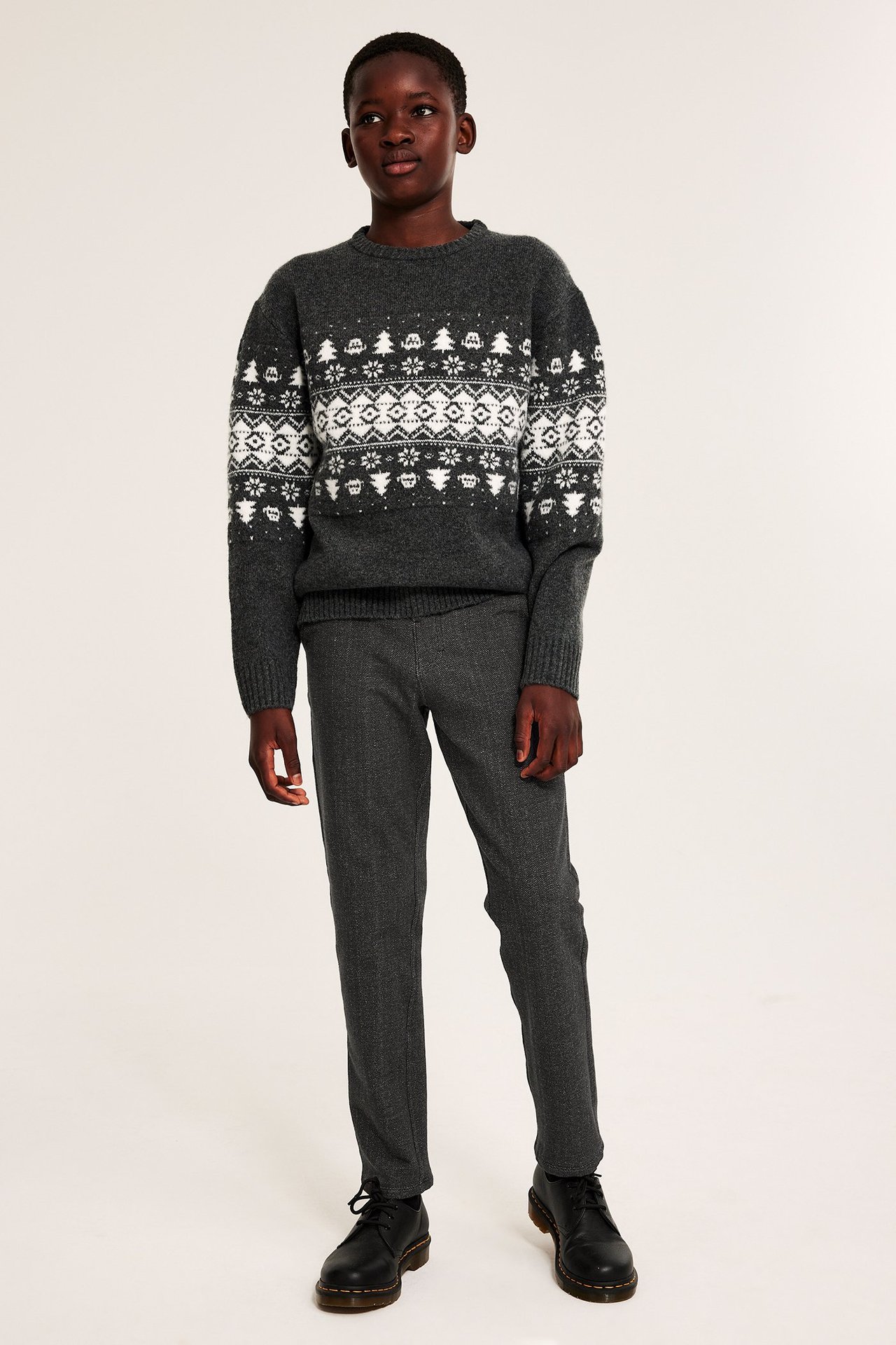 Edestäpäin lapsi tummanharmaissa kalanruotokuvioisissa housuissa, harmaassa fair isle -neuleessa ja mustissa nauhakengissä.