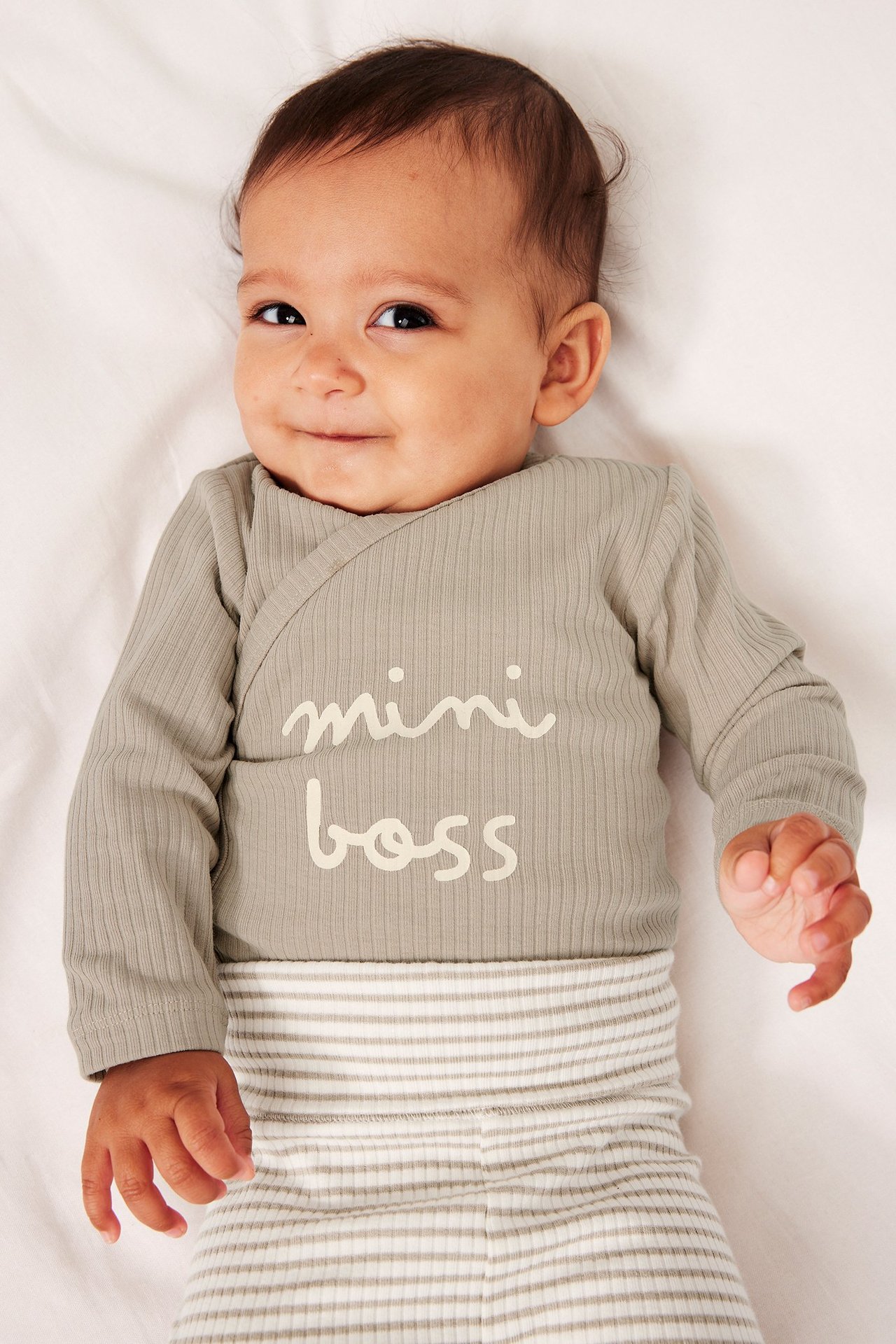 Hymyilevä vauva beigessä ribbineulotussa "mini boss" bodyssä ja raidallisissa housuissa, edestäpäin.