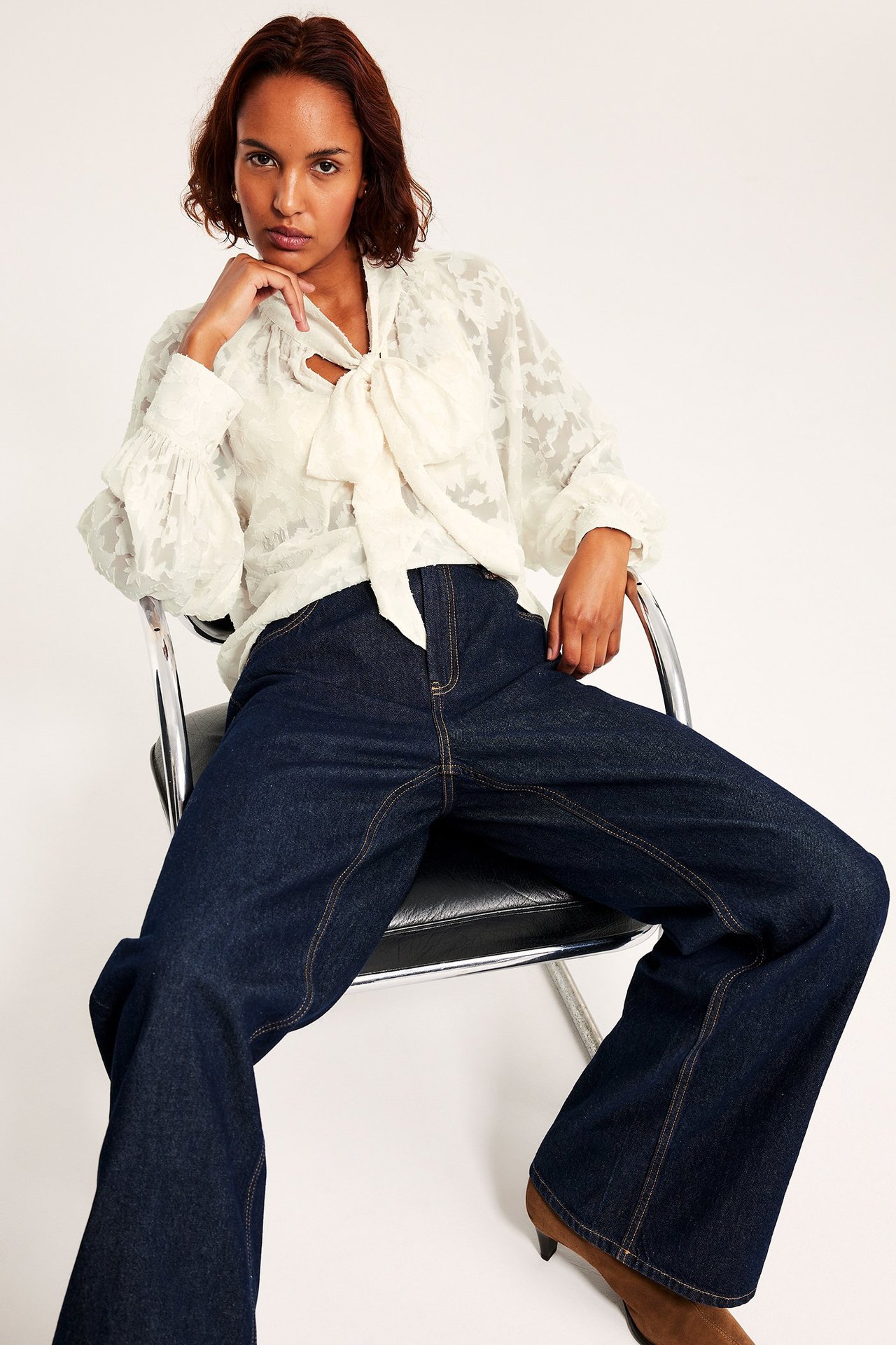 Kobieta w koronkowej bluzce w kolorze ecru, szerokie jeansy, siedzi na krześle.