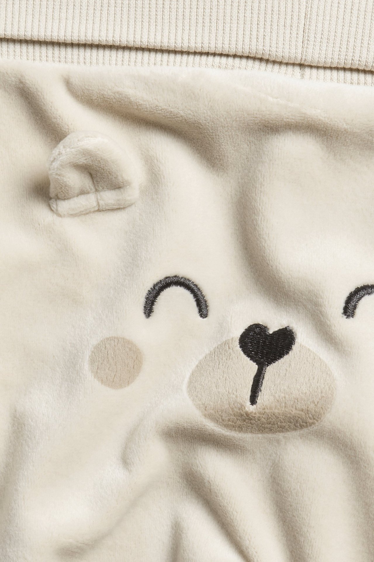 Zbliżenie beżowych welurowych spodni dresowych dla niemowląt z haftowaną twarzą misia i uszami.