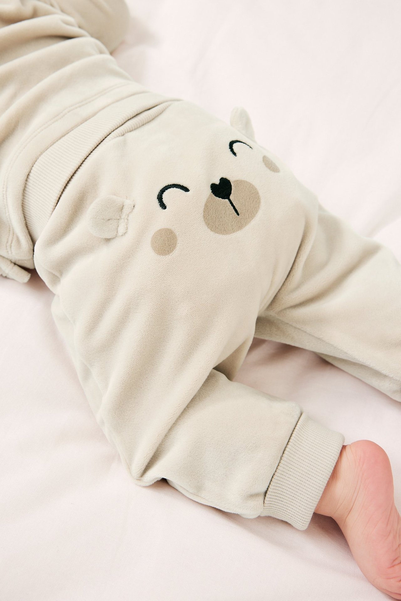 Lähikuva vauvasta beigenvärisissä velour-collegehousuissa, joissa on karhuapplikaatio ja yhteensopiva paita.