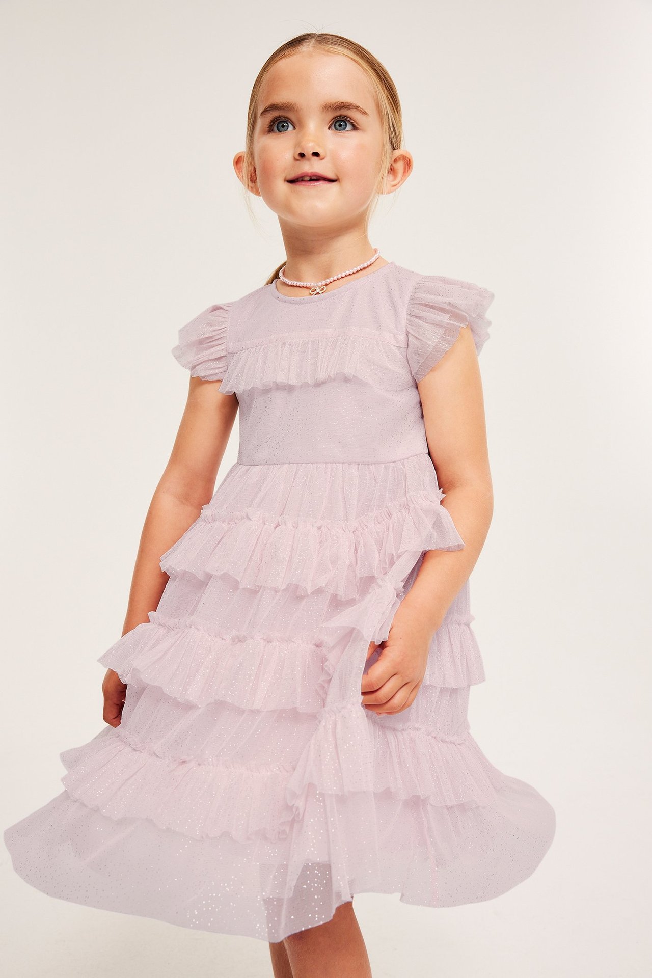 Framsida: Barn i en ljusrosa glittrig tyllklänning med volanger och ett rosa pärlhalsband.