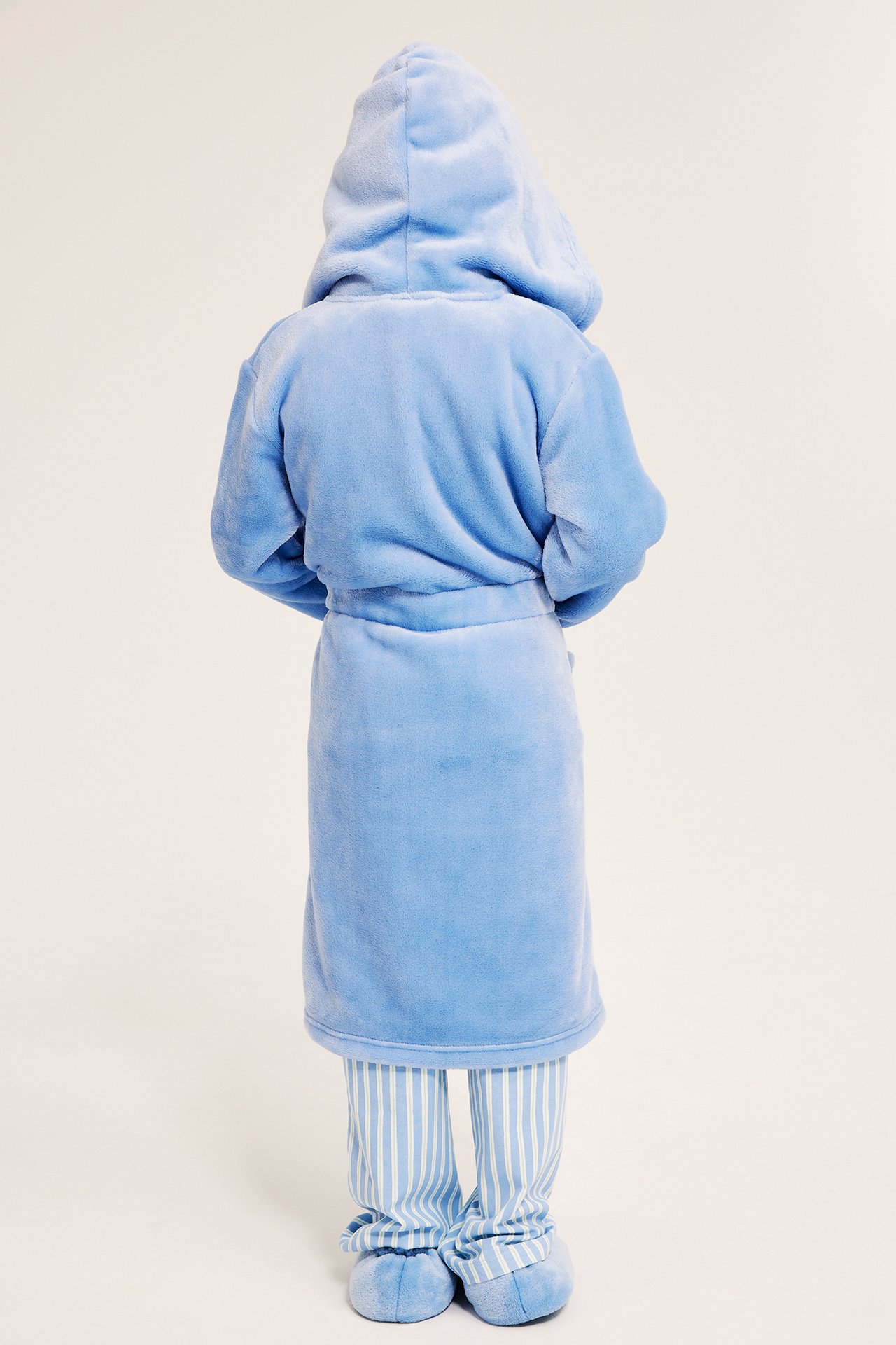 Bakfra, et barn i en lys blå hettefleece-morgenkåpe over stripete pyjamas og blå tøfler.