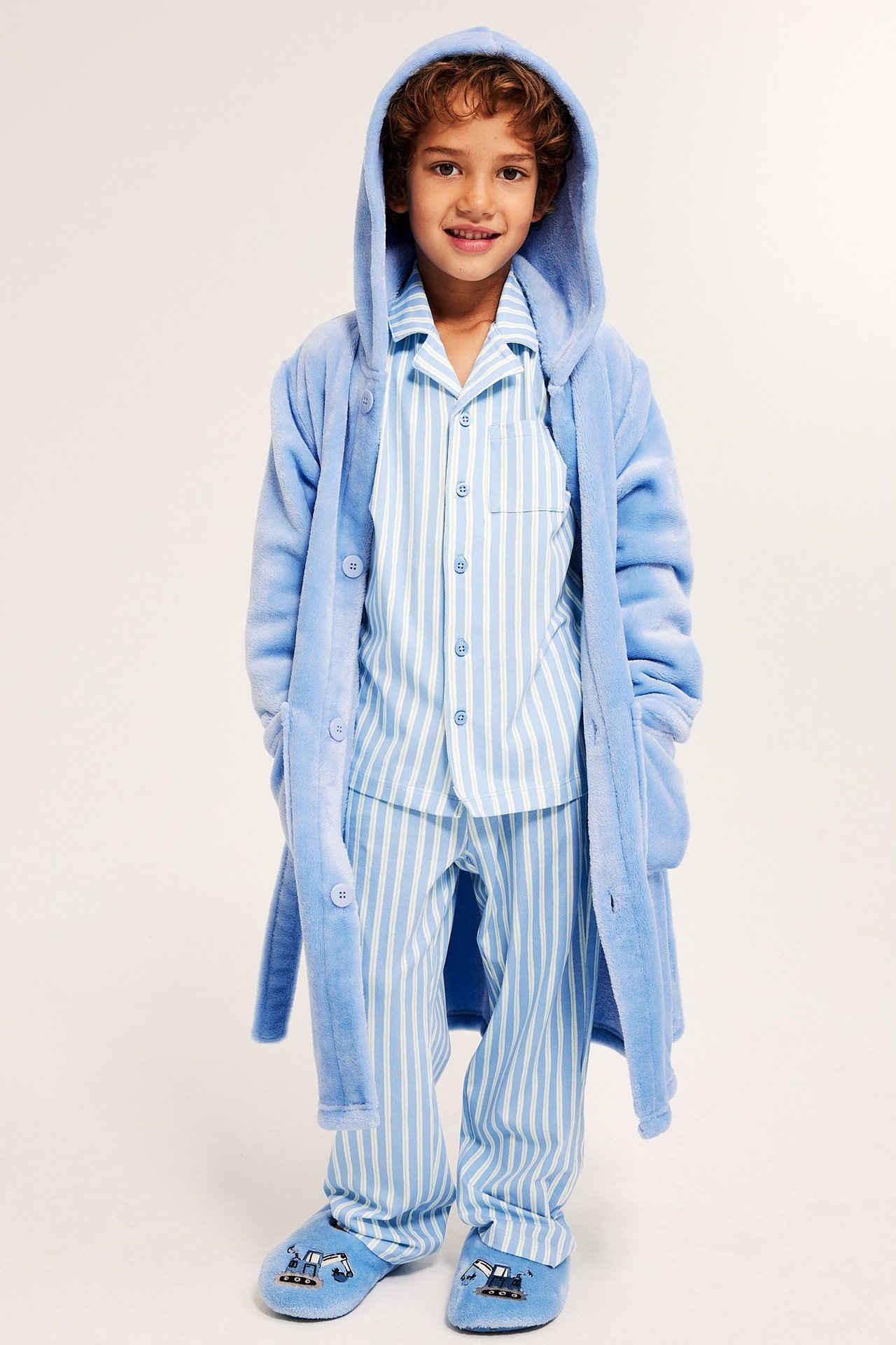 Edestä kuvassa lapsi vaaleansinisessä hupullisessa fleece-kylpytakissa, raidalliset pyjamat ja kaivuritossut.