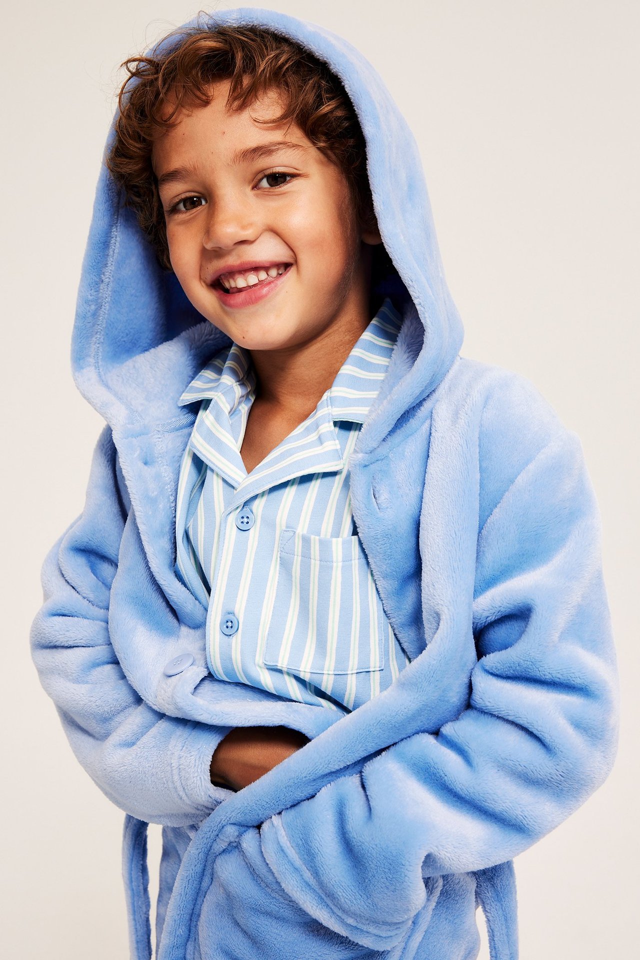 Edestäpäin hymyilevä lapsi vaaleansinisessä hupullisessa fleecerokissa raidallisen pyjaman päällä.