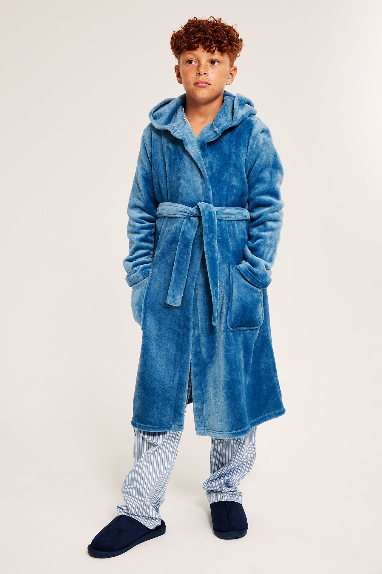 Edestäpäin: Lapsi sinisessä hupullisessa fleece-aamutakissa vyöllä, raidallisessa pyjamassa ja laivastonsinisissä tohveleissa.