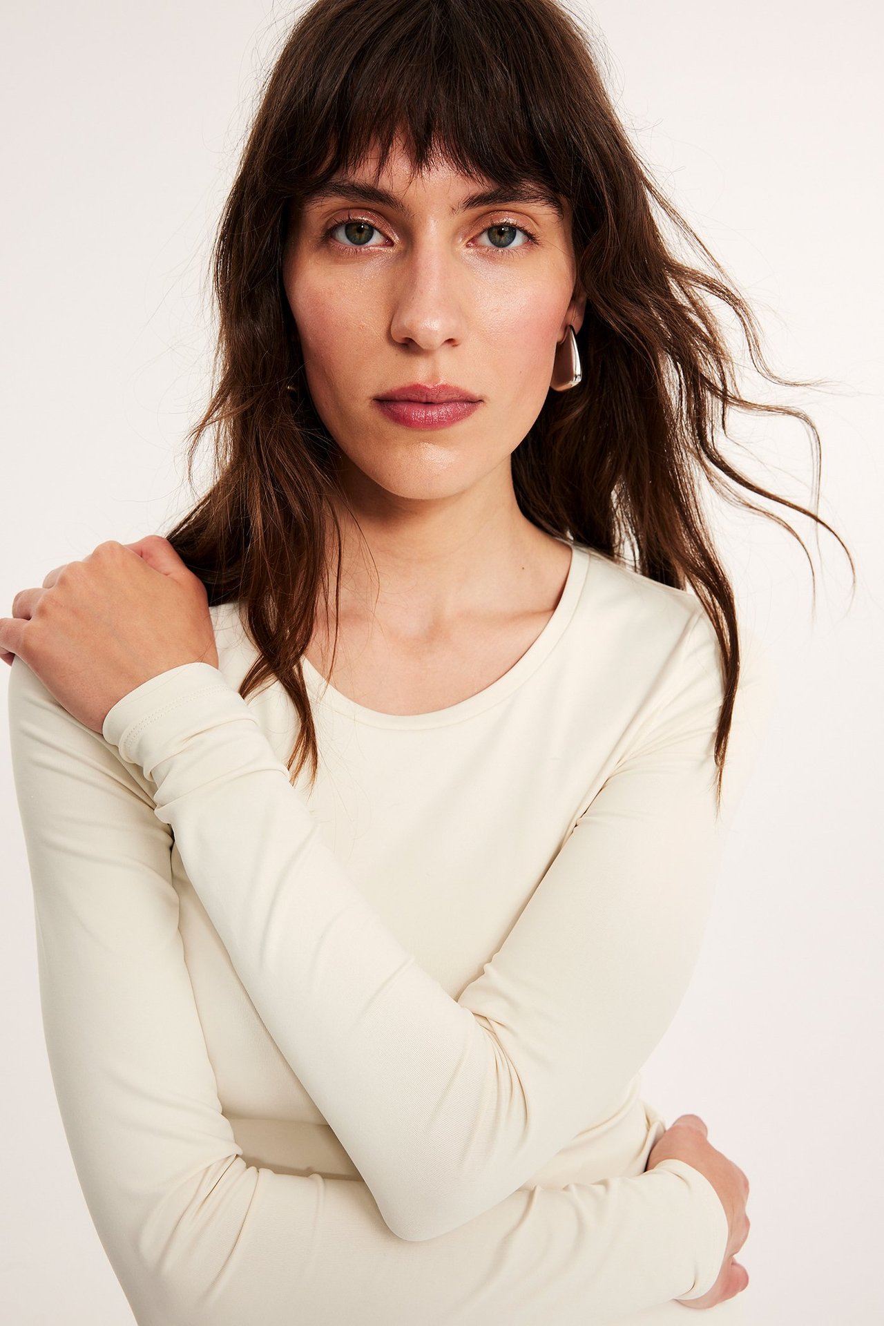 Närbild framifrån på en kvinna i en offwhite långärmad topp med rund hals och silverfärgade örhängen.