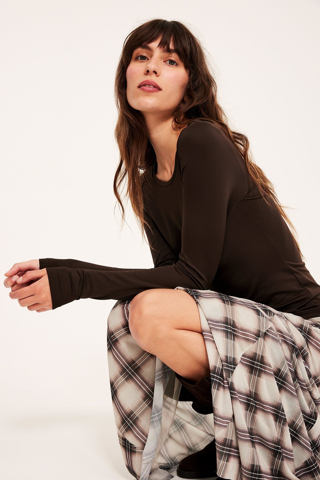 Forfra, kvinne i mørkebrun langermet rundhalset topp og et lyst rutete skjørt.