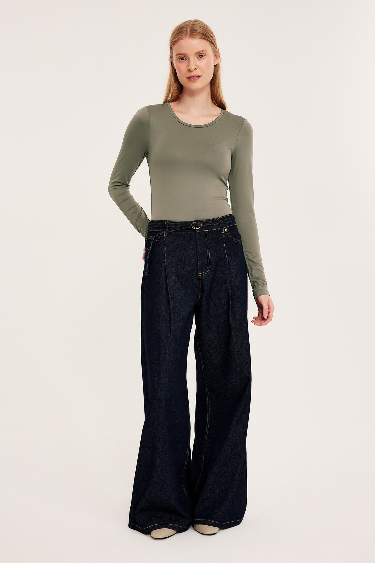 Forfra: kvinne i en figurnær brun langermet topp, mørkeblå vide jeans og lyse sko.