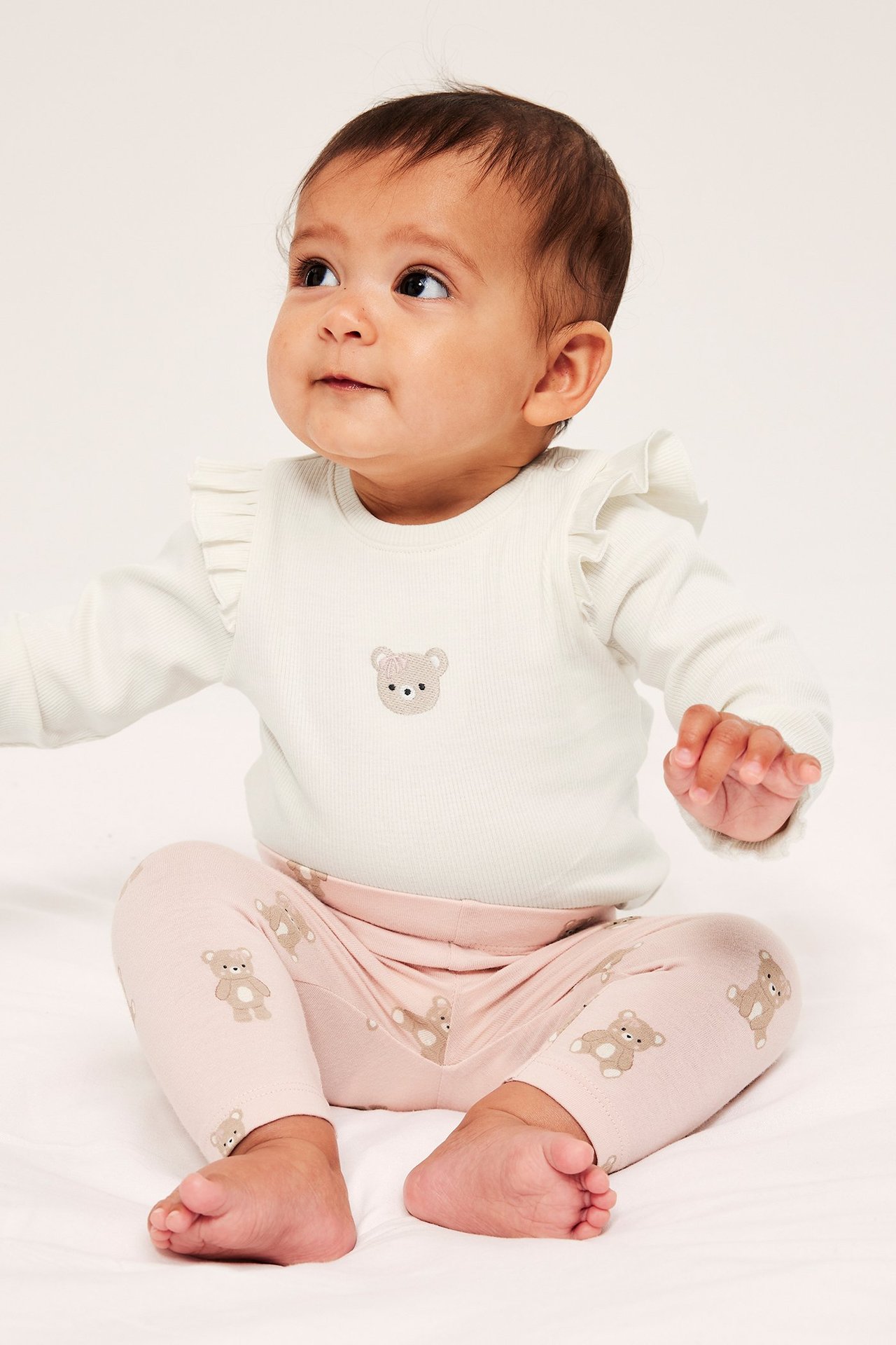 Edestä, vauva yllään luonnonvalkoinen ribattu body röyhelöillä ja nallekirjonnalla, sekä vaaleanpunaiset nallekuvioiset leggingsit.