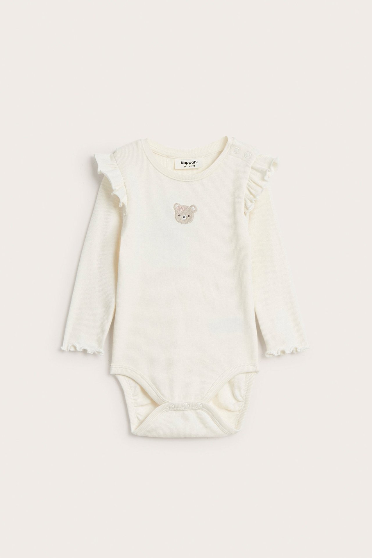 Edestä näkymä luonnonvalkoisesta ribatusta vauvan body-paidasta, jossa olkapäässä röyhelöt ja brodeerattu nalle.