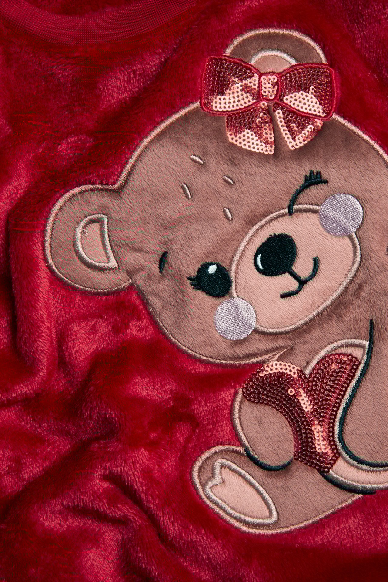 Zbliżenie czerwonej puszystej bluzy poliestrowej z haftowanym misiem, kokardą z cekinów i sercem.