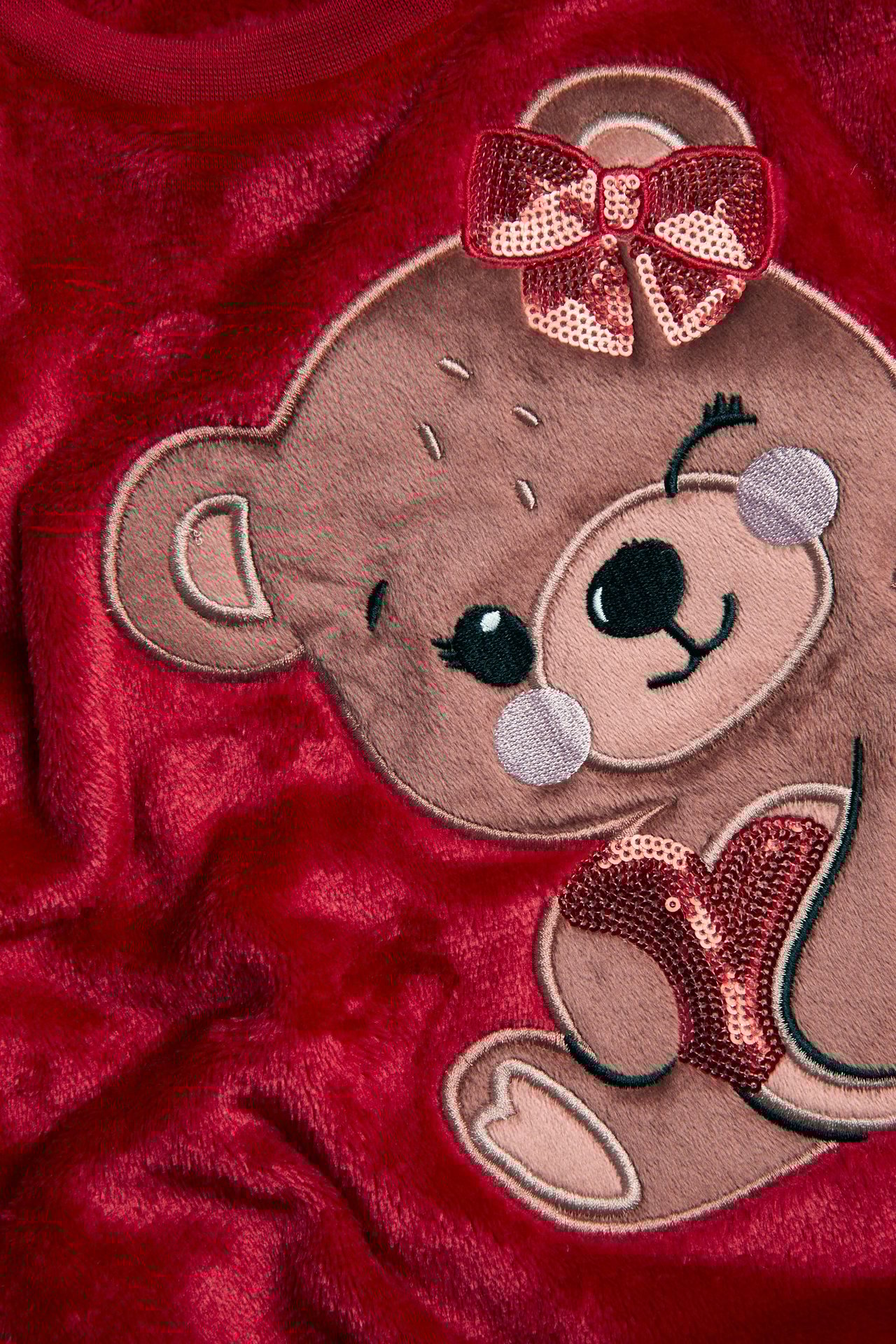 Zbliżenie czerwonej puszystej bluzy poliestrowej z haftowanym misiem, kokardą z cekinów i sercem.