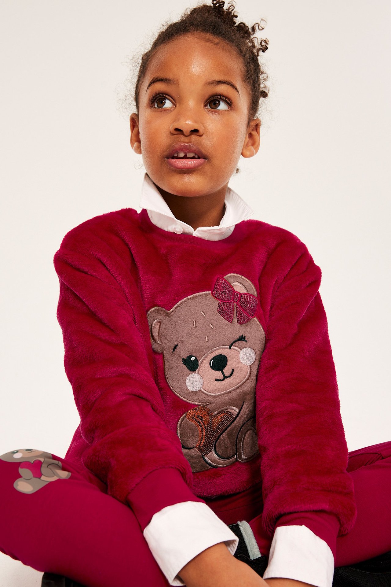 Edestä lapsi punaisessa pörröisessä nalleprinttipaidassa, valkoisen paidan ja punaisten housujen päällä.