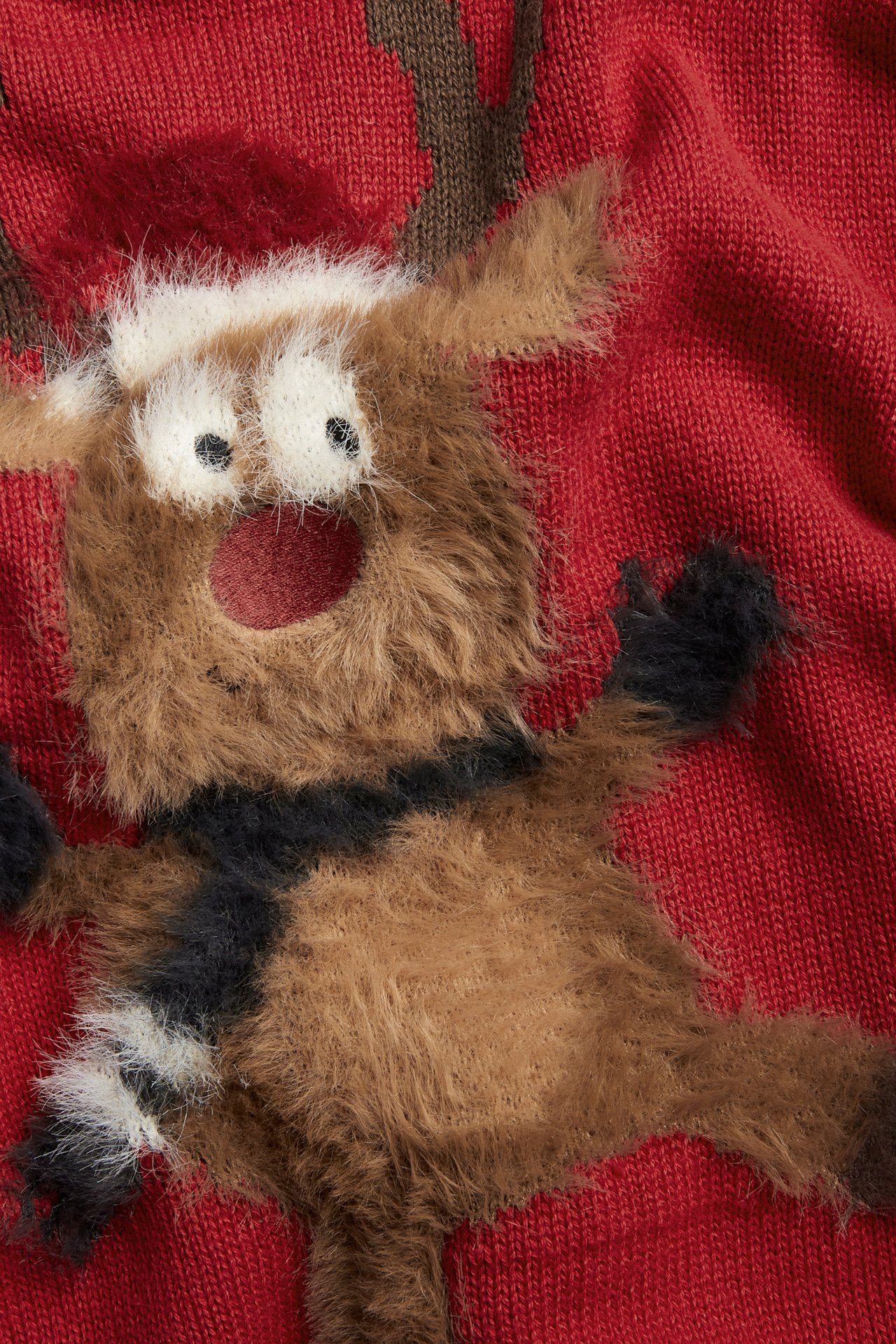 Närbild av en röd stickad jultröja för barn med en lurvig brun Rudolf-applikation.