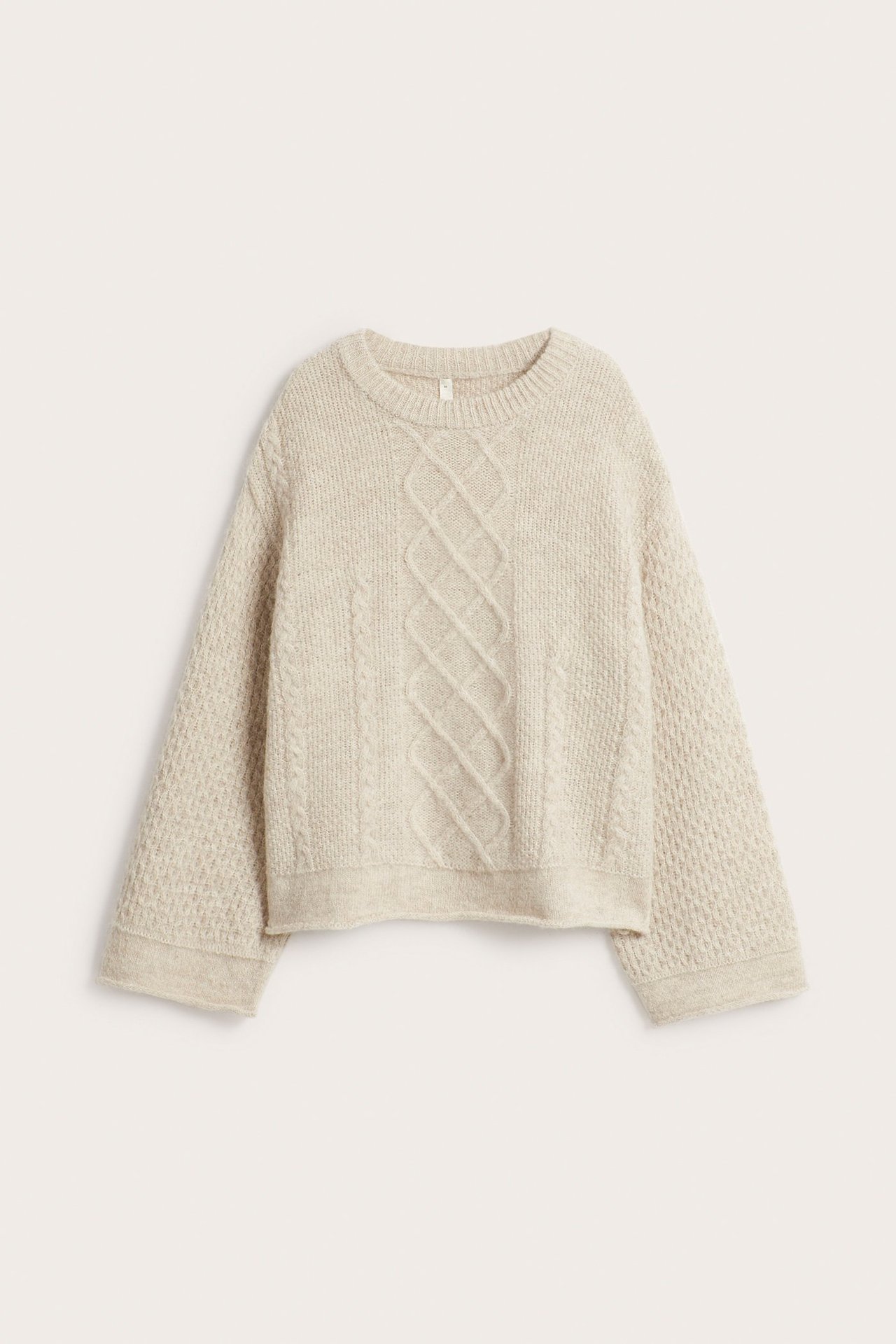 Frontvisning av en beige oversized strikkegenser i ullblanding med rund hals og flettestrikkede mønstre.