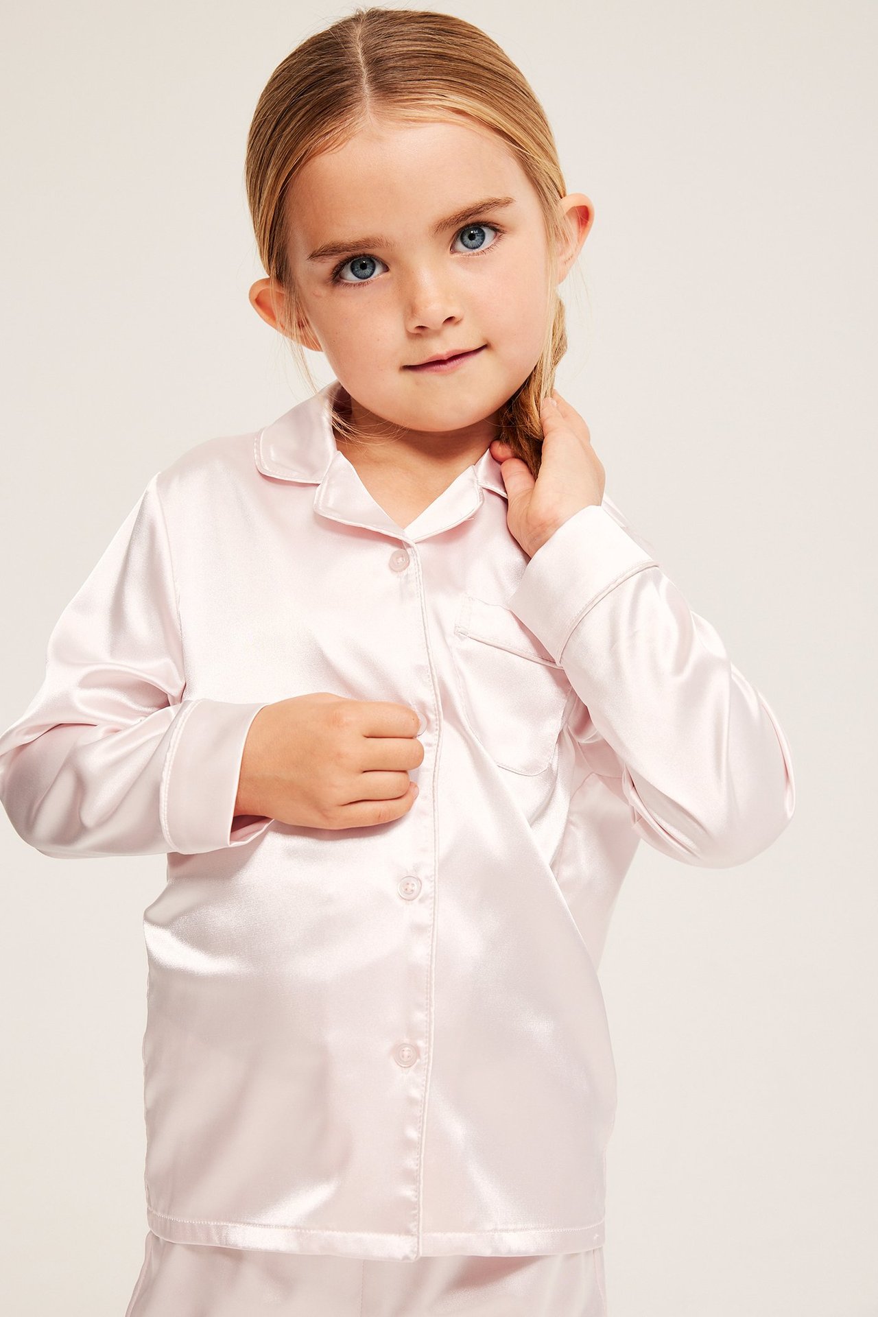 Edestä, lapsi vaaleanpunaisessa satiini pitkähihaisessa pyjamapaidassa napeilla. Yhteensopivat housut näkyvät.