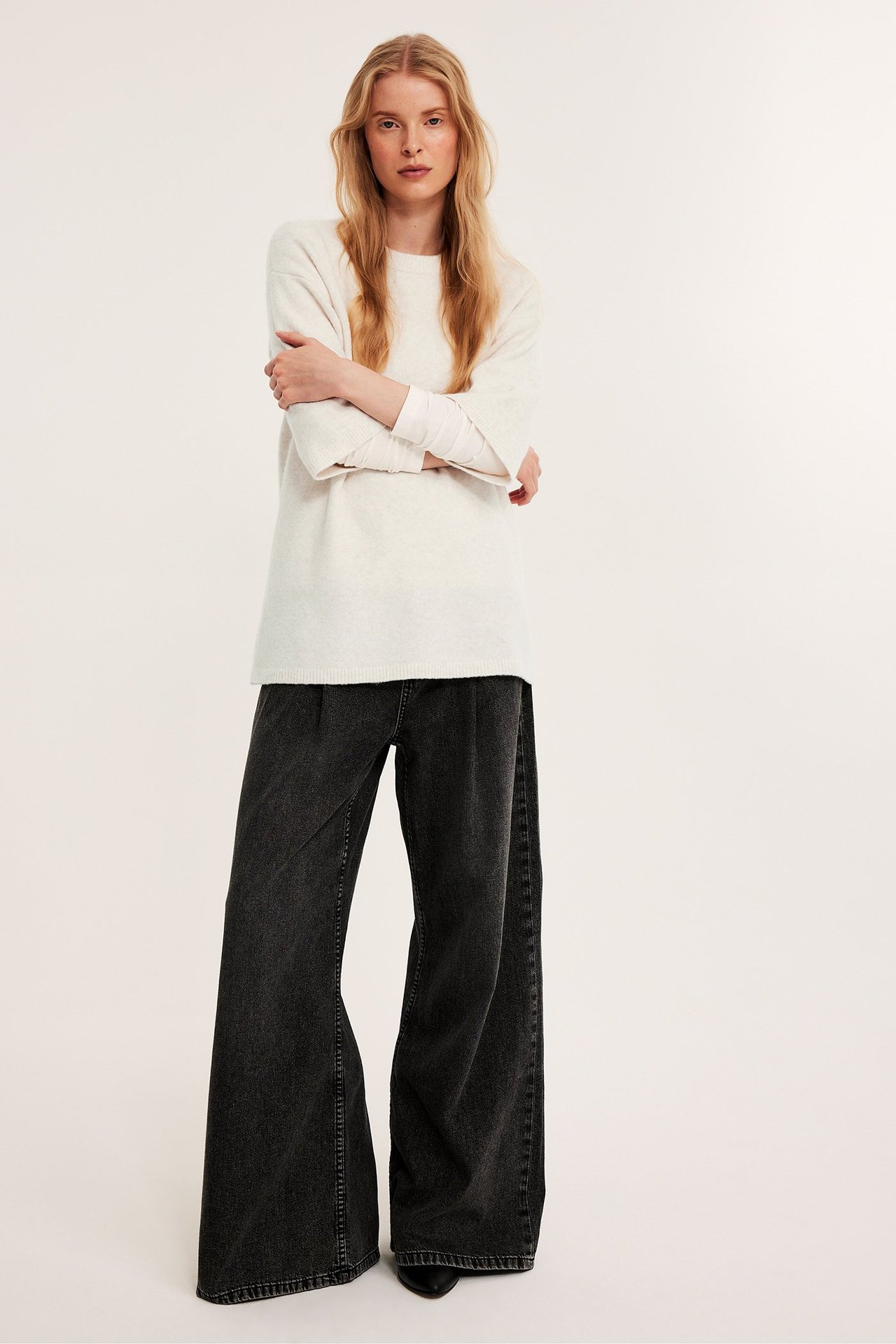 Forfra: Kvinne i en lys beige lang oversized strikkegenser og mørke vide jeans.