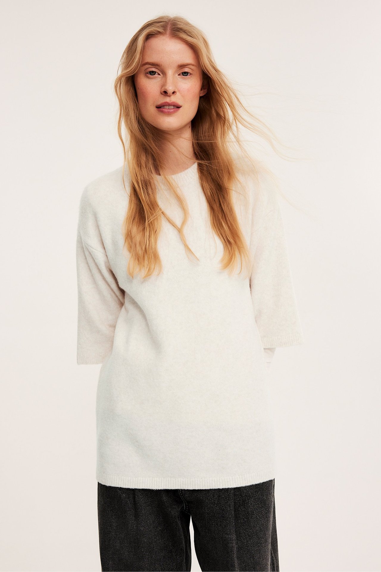 Widok z przodu: kobieta w długim, oversize'owym jasnobeżowym swetrze i ciemnoszarych spodniach.