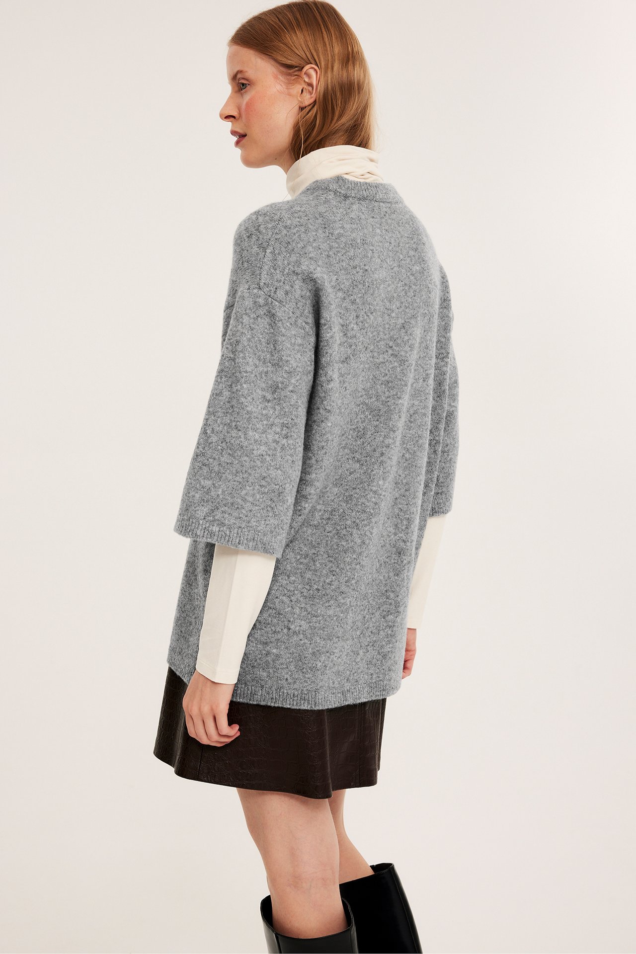 Sidebilde av kvinne i lys grå oversized strikket genser, kremfarget topp, brun skjørt, svarte støvler.