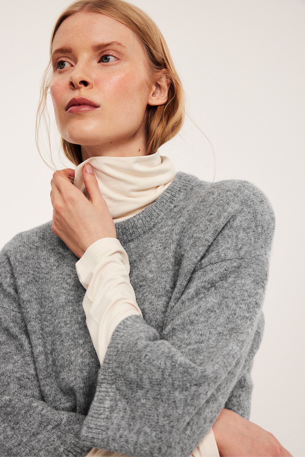 Nærbilde av en kvinne i en lys grå oversized strikkegenser over en lys beige høyhalset topp.