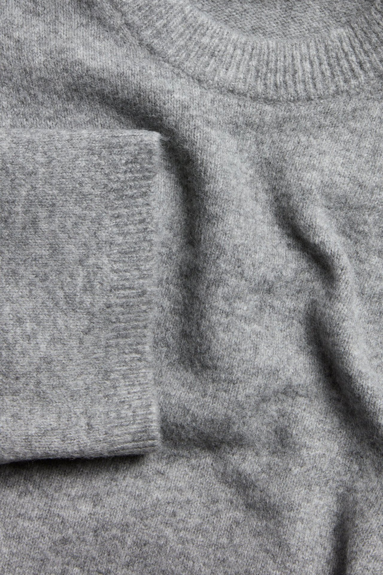 Zbliżenie jasnoszarego, długiego, oversize'owego swetra z dzianiny z prążkowanym okrągłym dekoltem i mankietem.