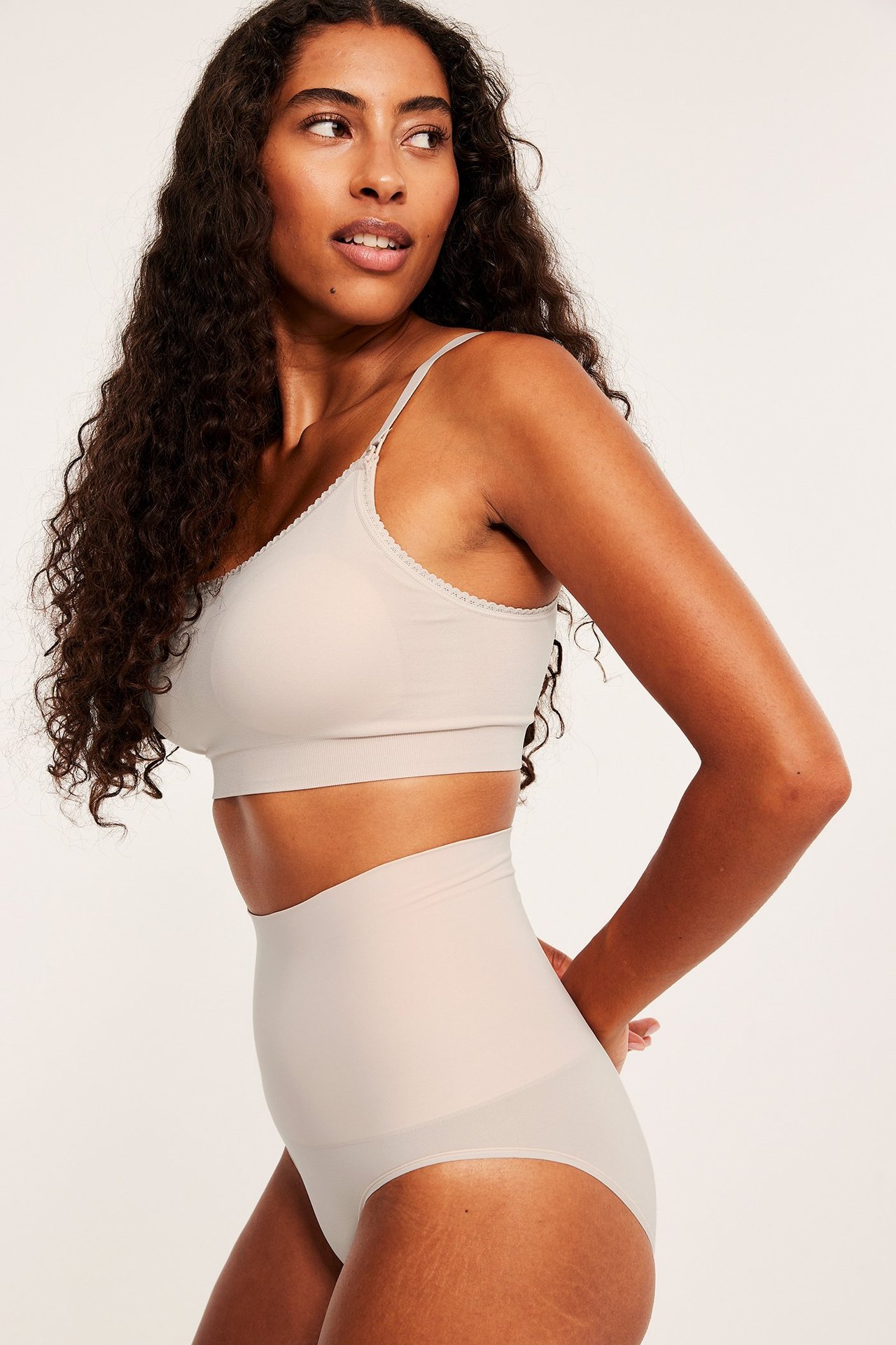 Sidebilde av en kvinne i lys rosa høy shapingtruse og matchende bralette med bølget kant.