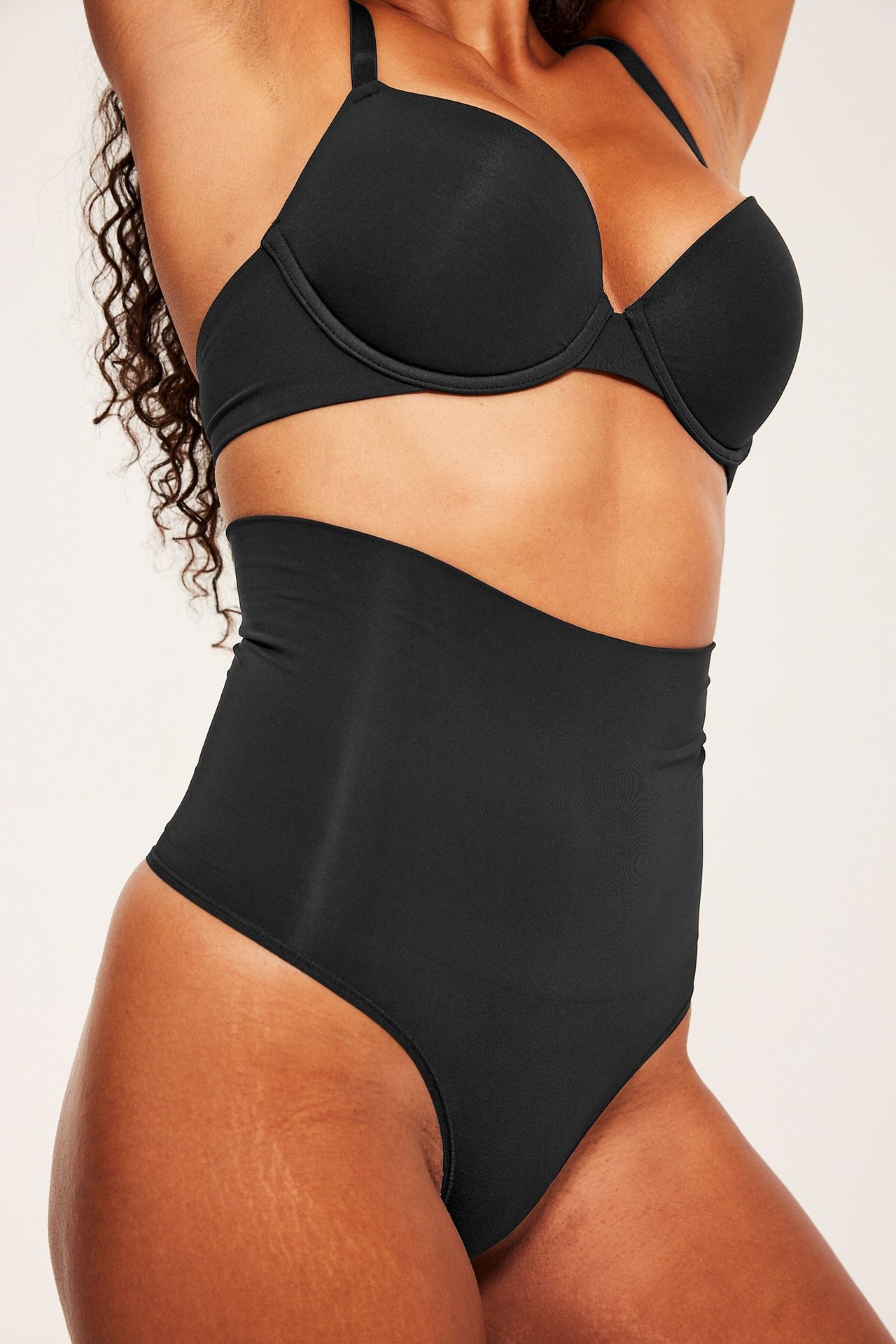 Frontvisning av en kvinne i svart høytlivs shape stringtruse og svart BH.