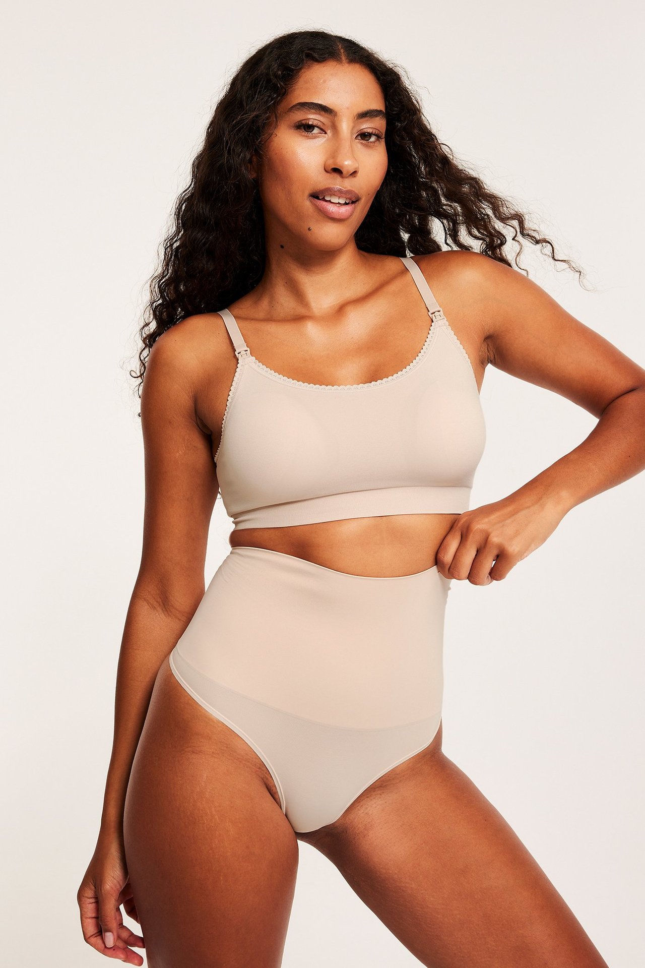 Edestä, nainen vaaleanpunaisissa korkeavyötäröisissä muotoilevissa stringeissä ja rintaliiveissä, vetää vyötärönauhaa.