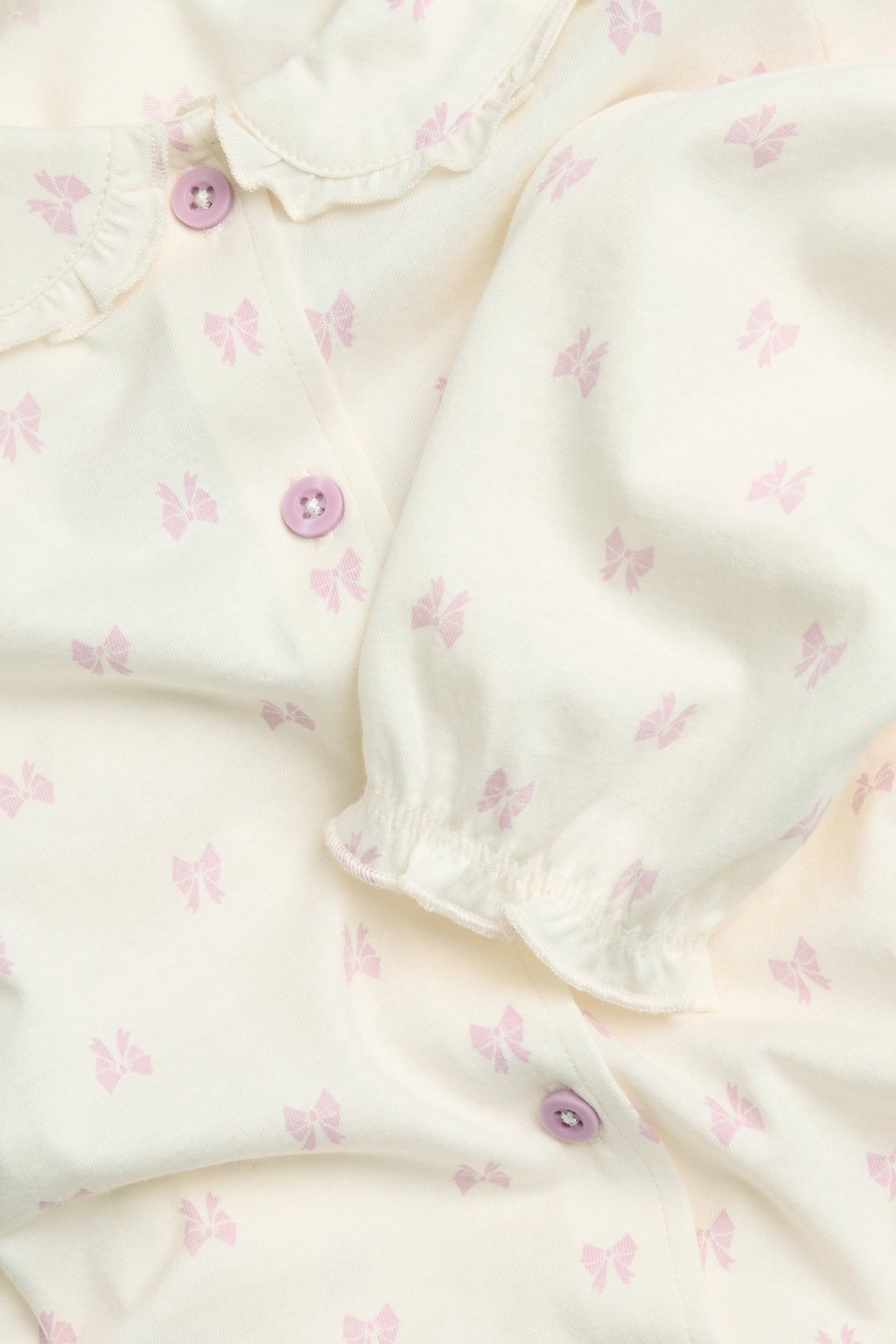 Zbliżenie: biała bawełniana piżama z różowymi kokardkami, falbankowym kołnierzem i liliowymi guzikami.