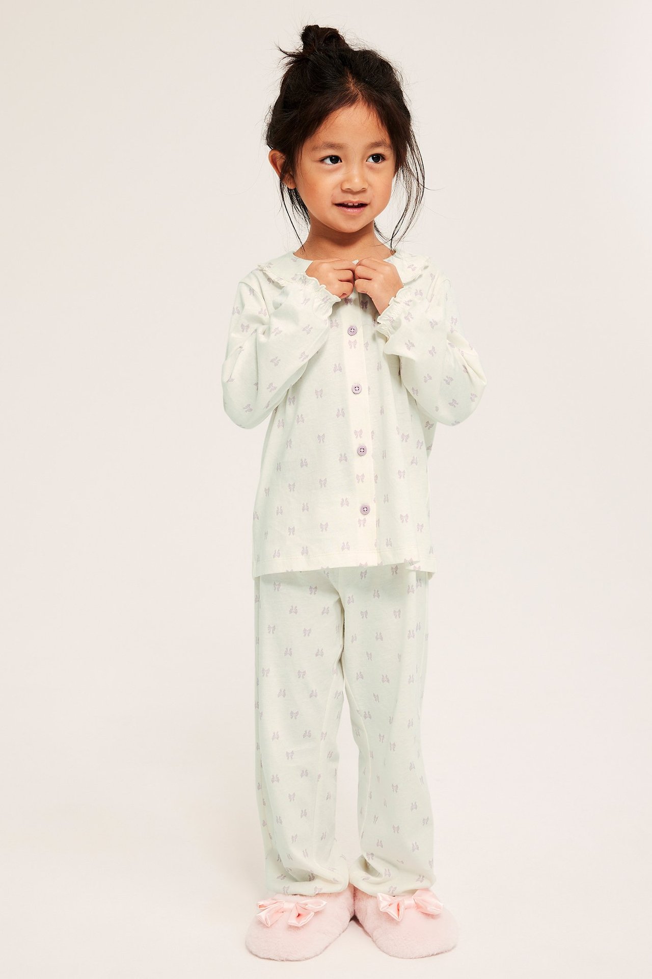 Widok z przodu: Dziecko w kremowej, wzorzystej piżamie bawełnianej z falbanowym kołnierzem, guzikami i różowymi kapciami.