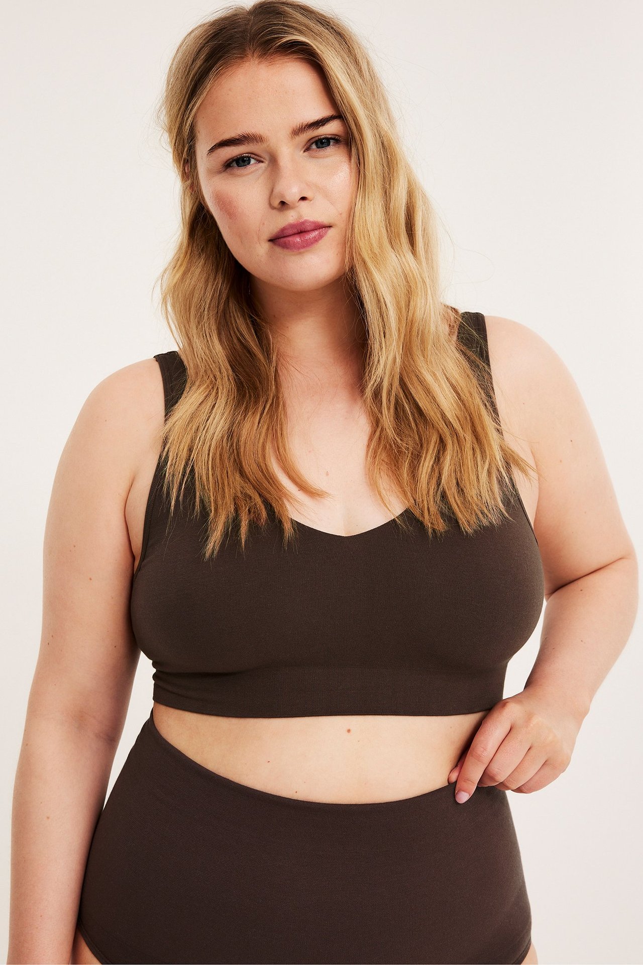 Kobieta w ciemnobrązowym bezszwowym biustonoszu z dekoltem w serek i figach, dłoń przy pasie.