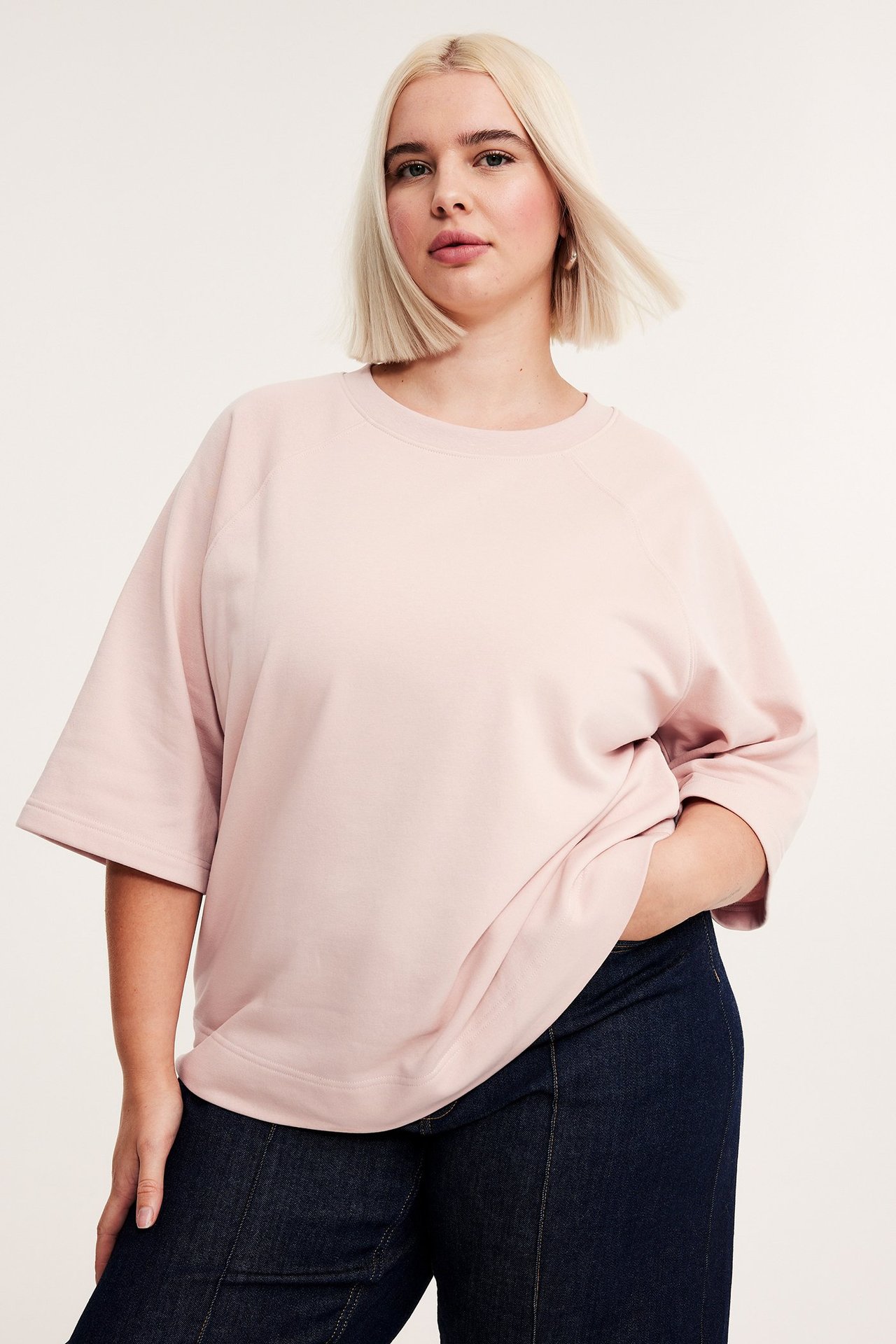 Widok z przodu: kobieta w luźnej, jasnoróżowej bluzie z krótkim rękawem i ciemnoniebieskich spodniach.