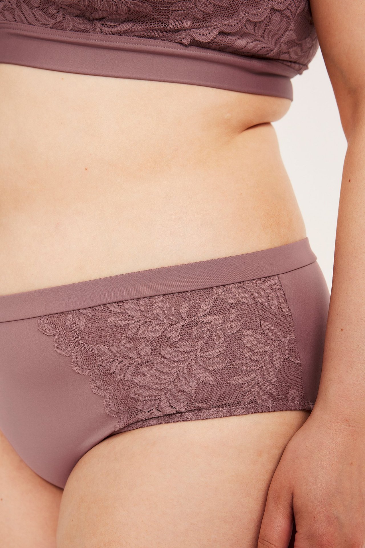 Lähikuva, sivukuva naisesta yllään tummanpunaiset mikrobriefit pitsiyksityiskohdilla ja yhteensopivat pitsirintaliivit.