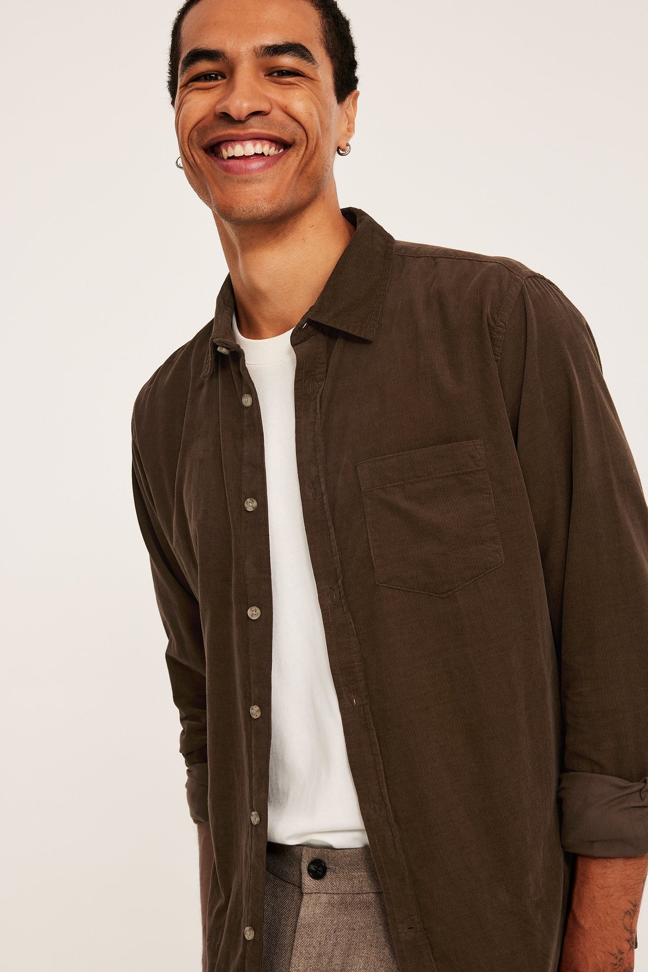 Edestä hymyilevä mies ruskeassa avoimessa vakosamettipaidassa valkoisen t-paidan ja vaaleanruskeiden housujen päällä.