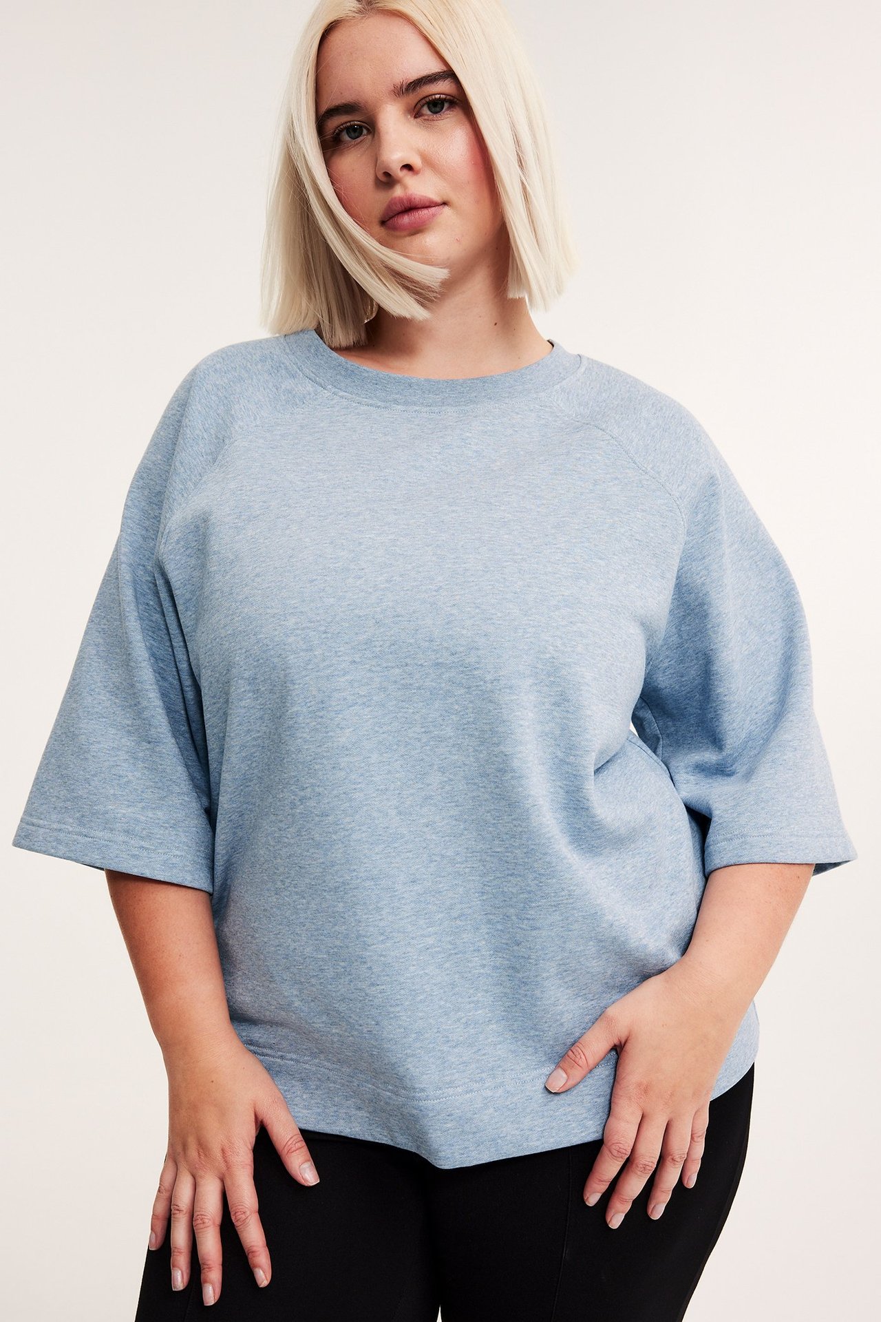 Widok z przodu kobiety w jasnoniebieskiej melanżowej bluzie z krótkim rękawem i czarnych spodniach.