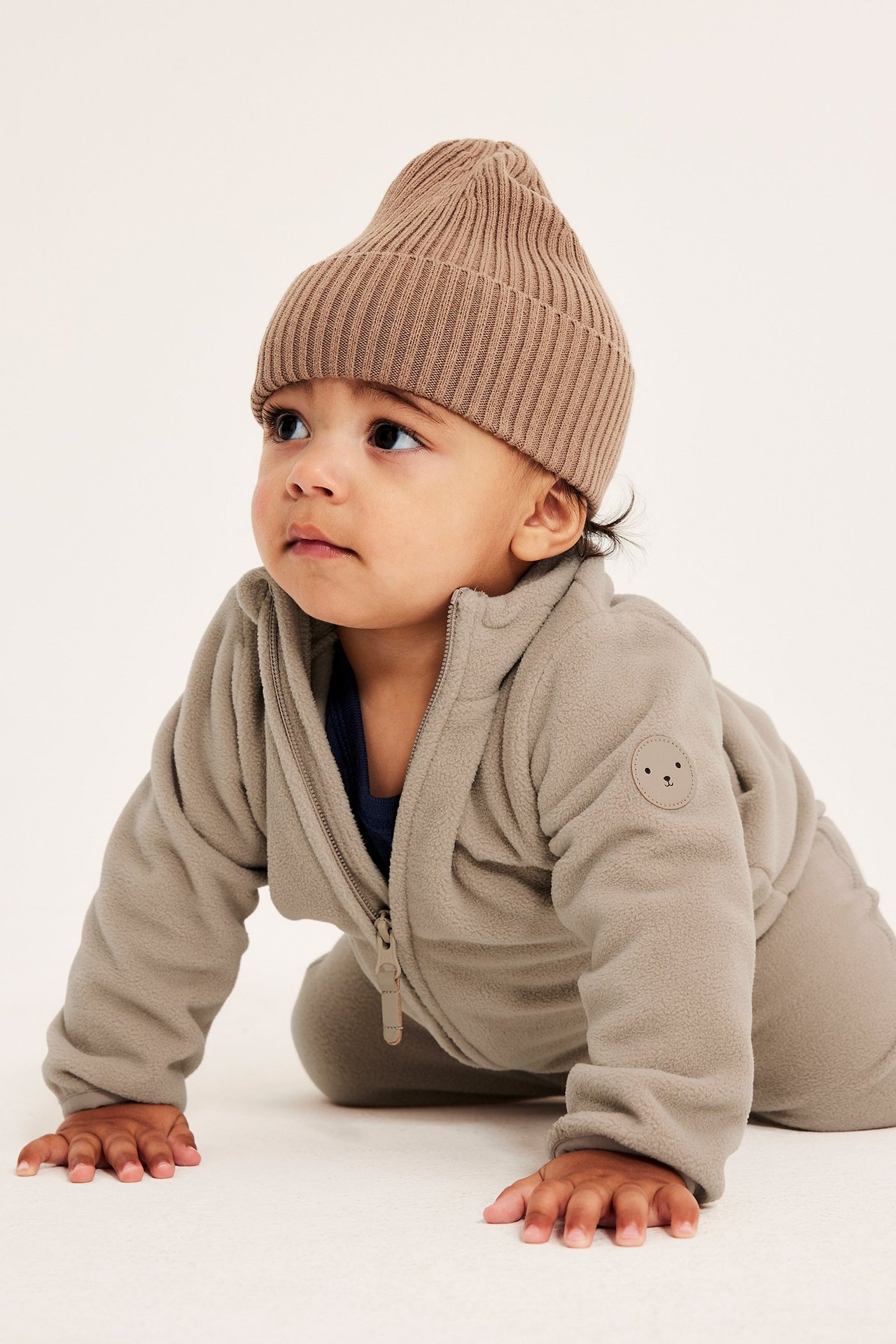 Forfra: krypende baby i beige fleeceoverall med glidelås og merke, samt brun ribbestrikket lue.