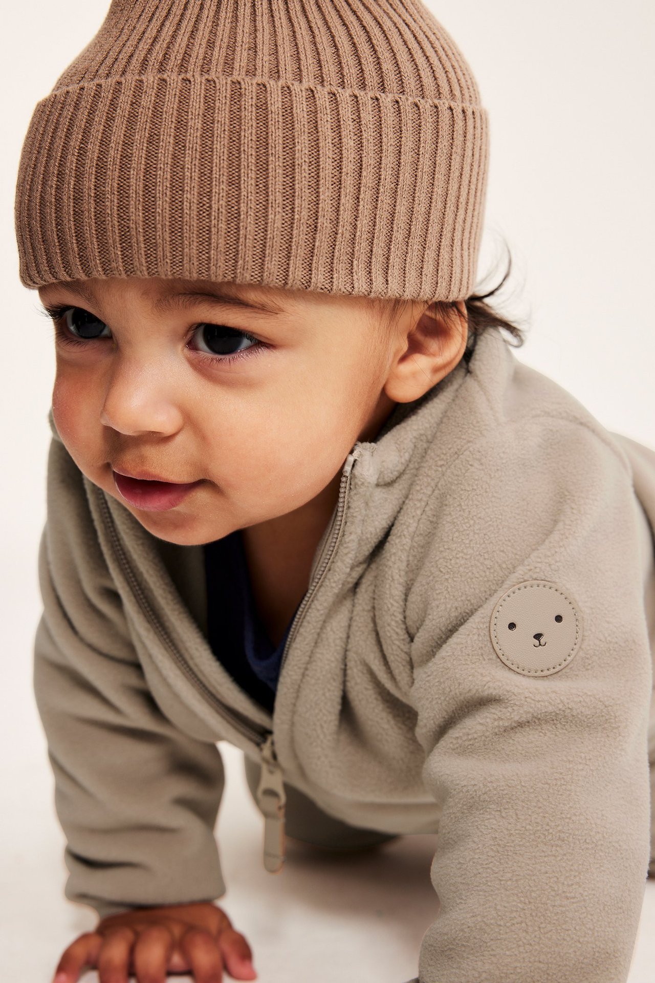 Forfra, baby i beige fleecejakke med bjørnemerke og brun ribbestrikket lue.