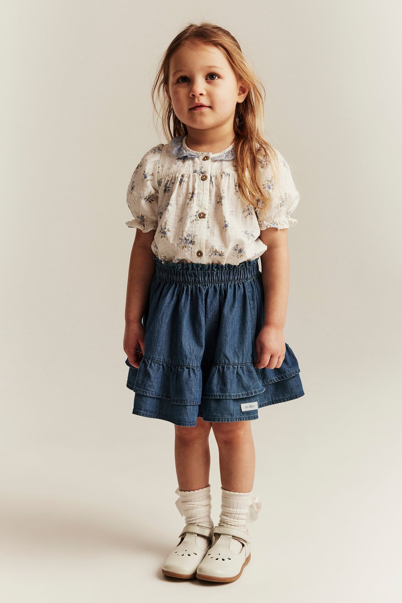 Framifrån: Kid i blå volangkjol i denim, vit blommig blus, vita strumpor och vita skor.