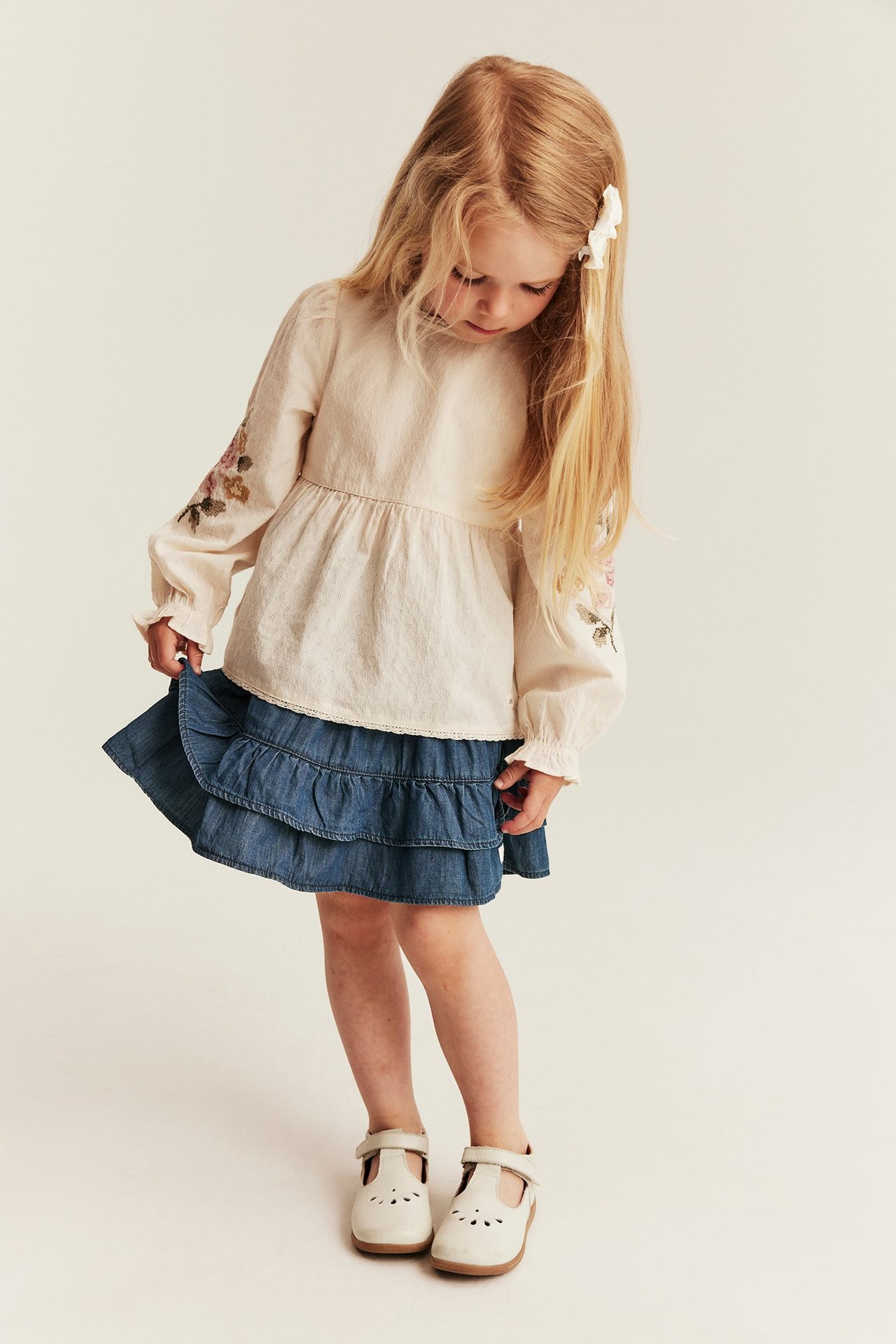 Forfra: Barn i blå denimskjørt med volanger, beige brodert bluse, hvit hårspenne og T-stroppesko.