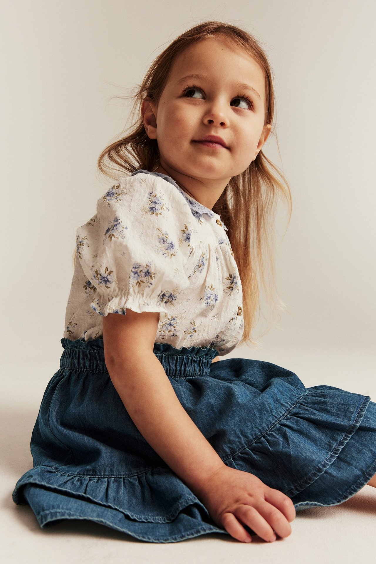Siedzące dziecko z boku w niebieskiej spódnicy jeansowej z falbanką i białej bluzce w kwiaty.