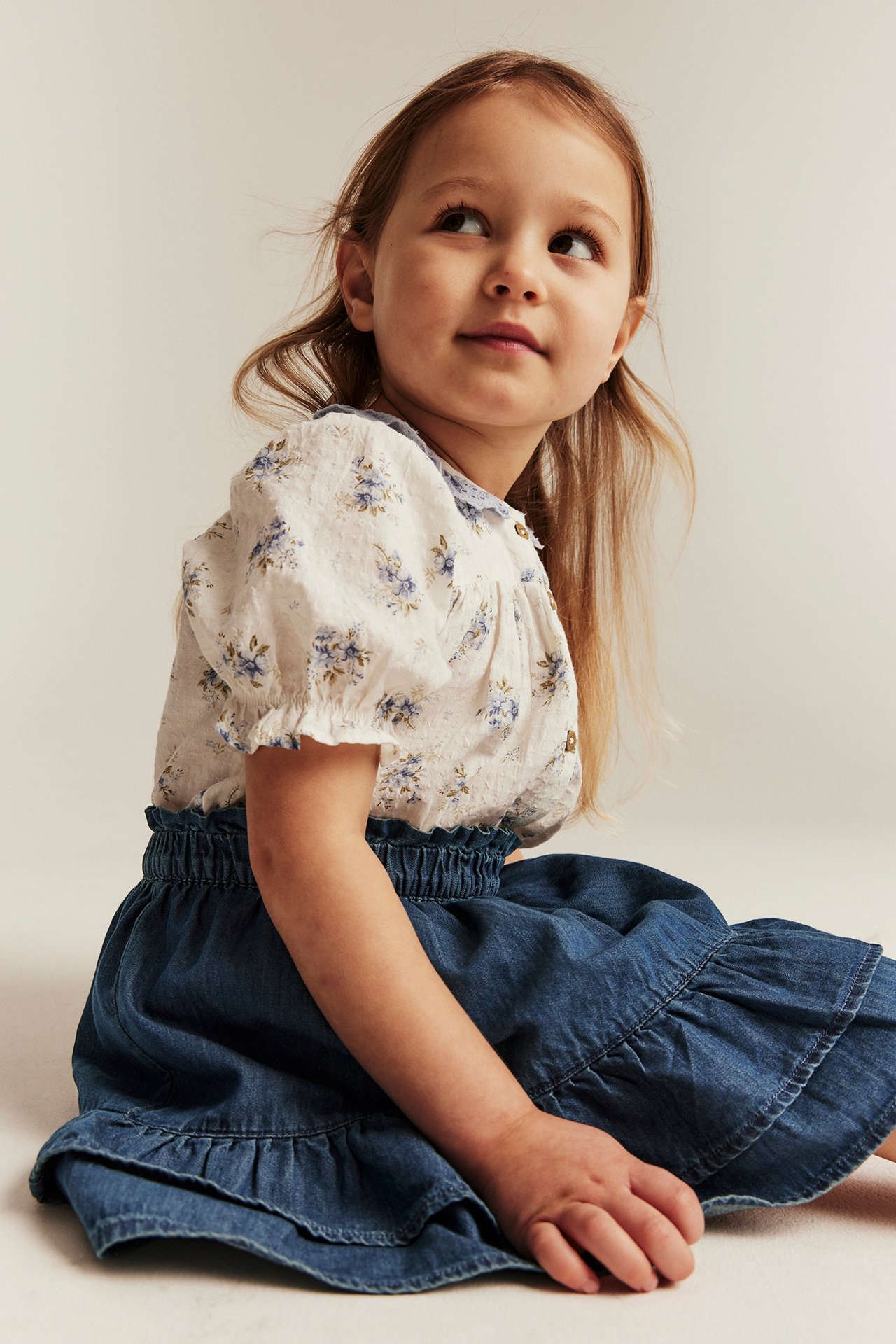 Framifrån, ett sittande barn i en blå denimkjol med volang och en krämvit blommönstrad blus.