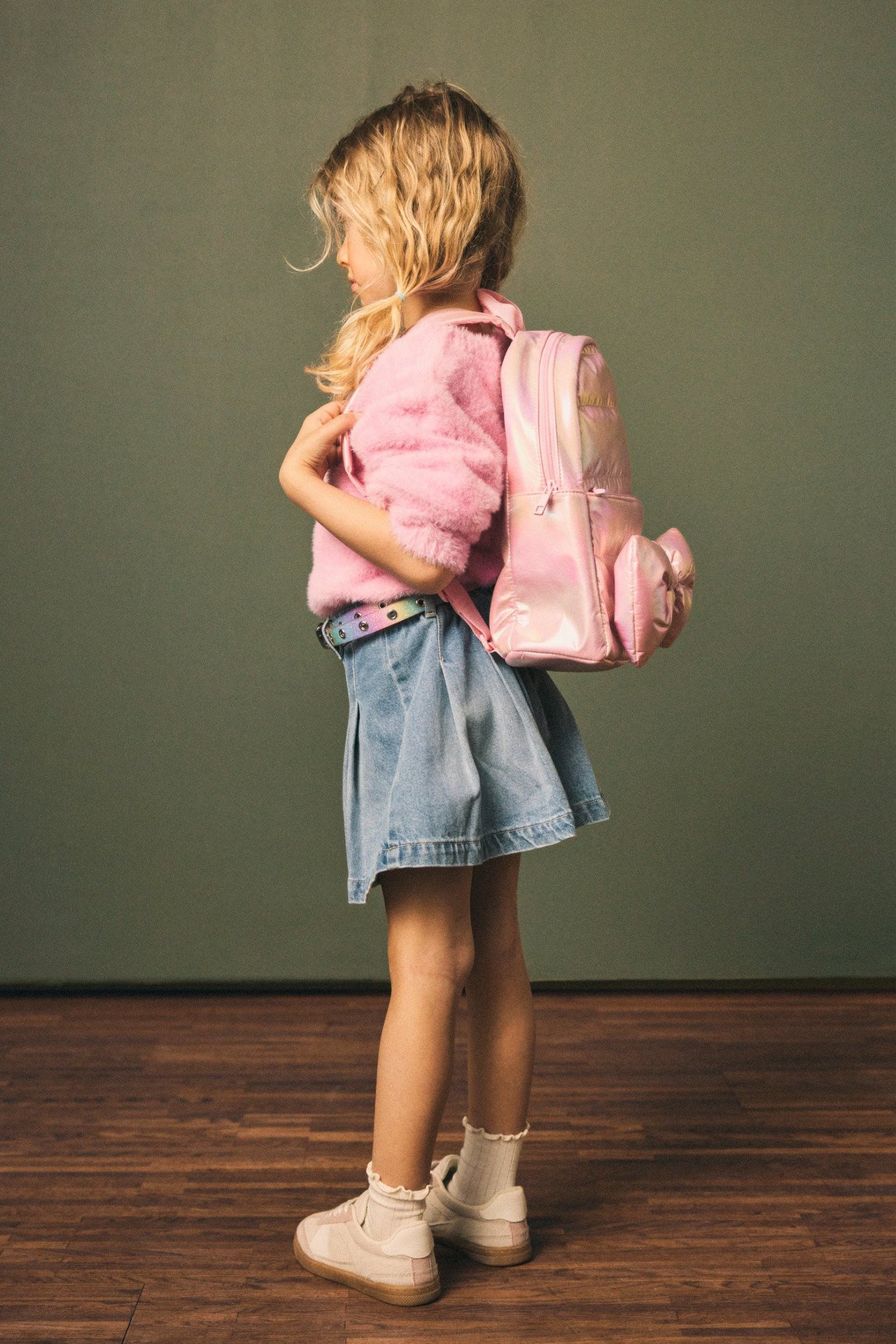 Sidovy av ett barn som bär en glänsande rosa ryggsäck med rosett, fluffig rosa tröja, jeanskjol och sneakers.