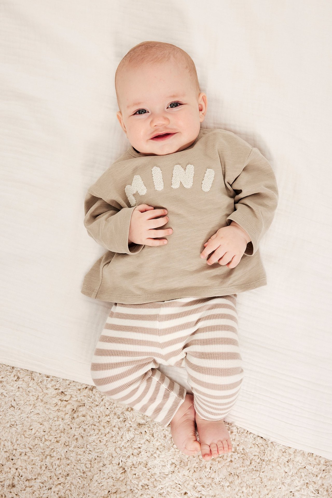 Widok z góry uśmiechniętego dziecka w beżowej koszulce z długim rękawem 'MINI' i legginsach w paski.